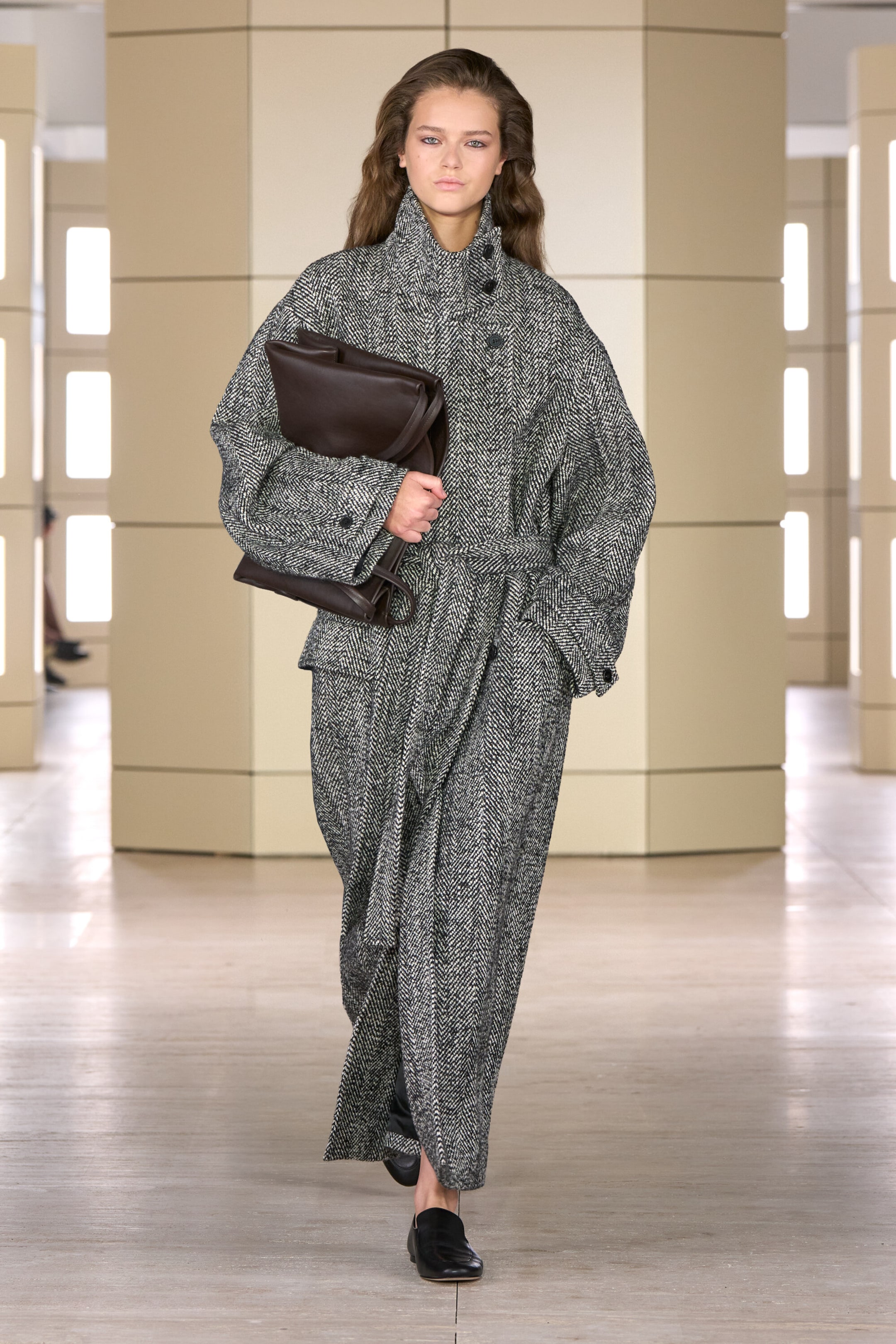 A model is wearing the belted herringbone wool funnel-neck coat in a front-facing view, showcasing its oversized, ankle-length silhouette and high buttoned funnel neck. The coat features a matching fabric belt cinched at the waist, wide dropped-shoulder sleeves, and is styled with a dark brown leather bag tucked under the arm. A model is wearing the belted herringbone wool funnel-neck coat in a front-facing view, showcasing its oversized, ankle-length silhouette and high buttoned funnel neck. The coat features a matching fabric belt cinched at the waist, wide dropped-shoulder sleeves, and is styled with a dark brown leather bag tucked under the arm.