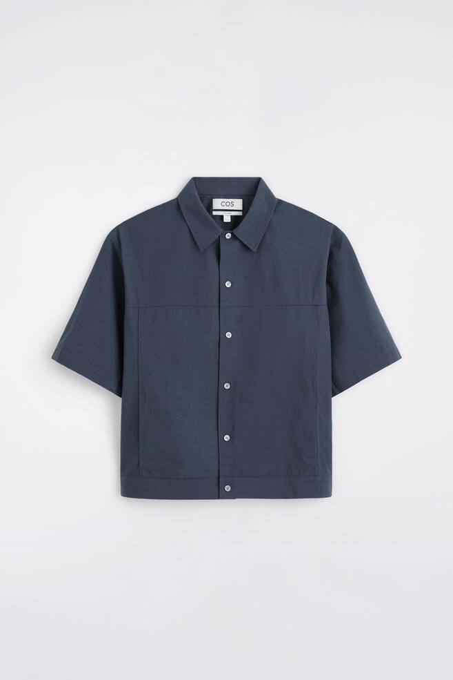 A dark blue men's shirt is facing front, featuring a collar and short sleeves. It is cut in a boxy shape, showcasing a button fastening with white buttons and visible topstitching forming a horizontal panel across the chest and vertical panels down the sides. - 1