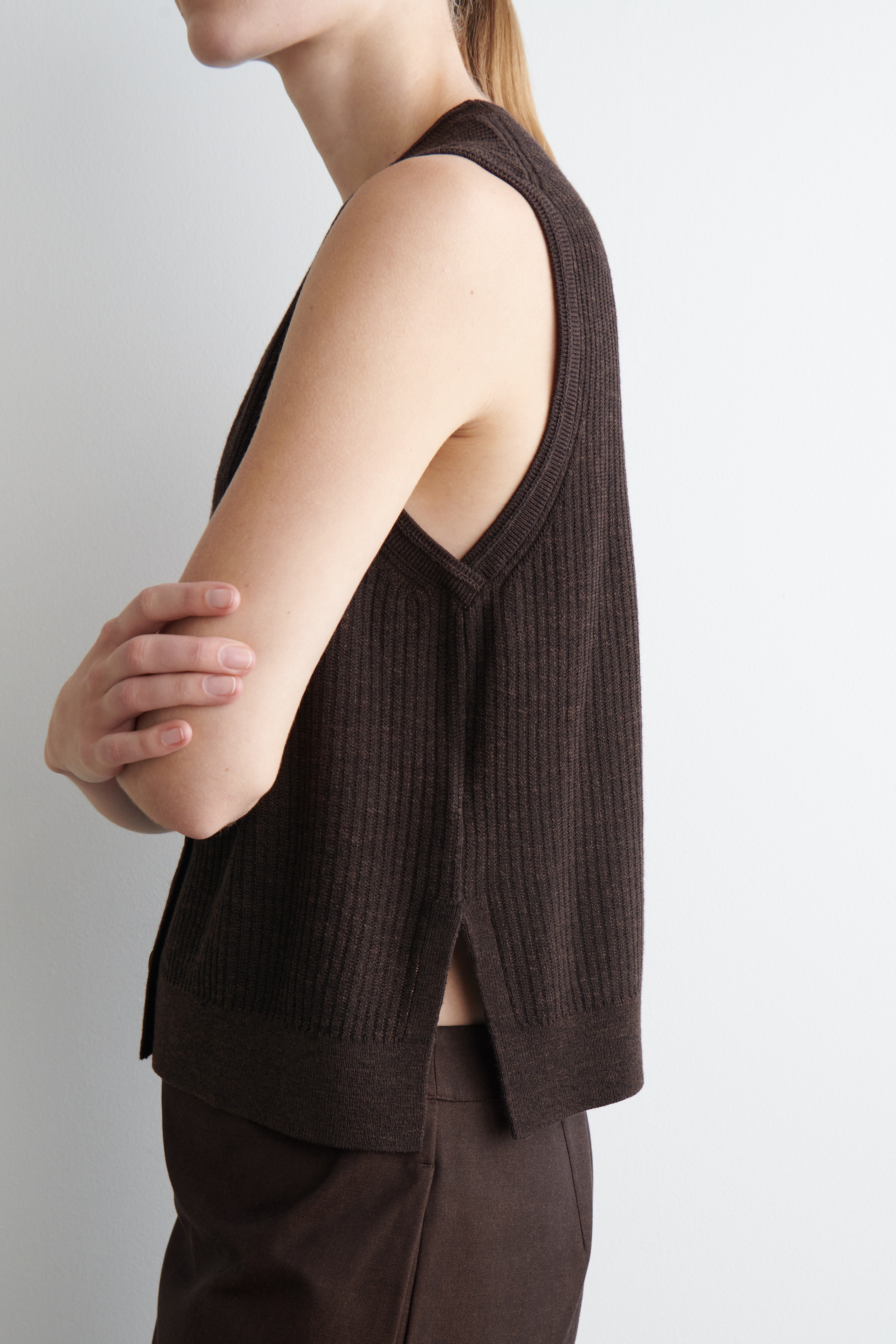 Ingrandisci l'immagine: Un gilet a maglia a coste marrone scuro viene mostrato angolato a sinistra, evidenziando il suo design senza maniche, scollo a V e spacchi laterali all'orlo.