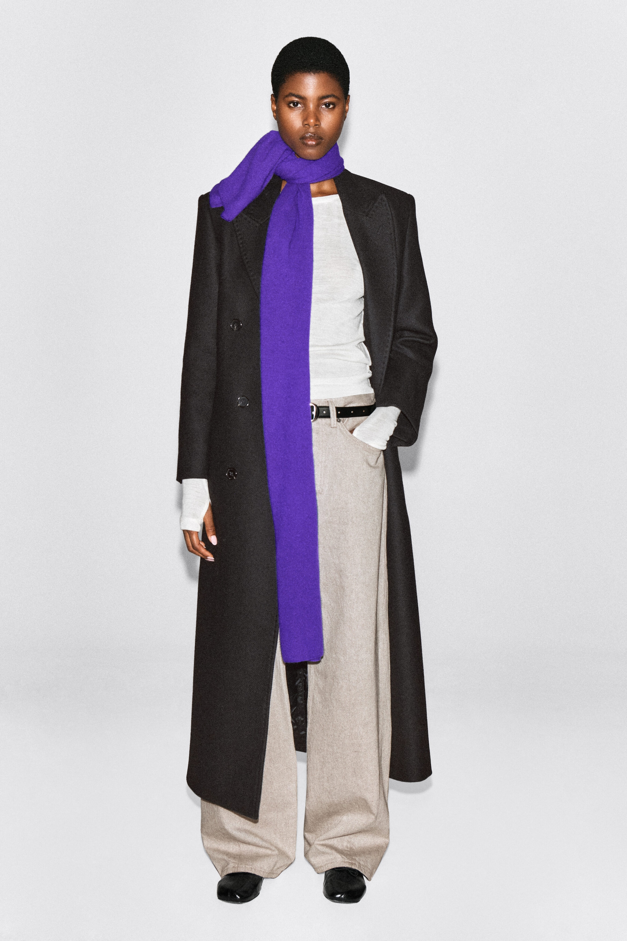 The model is wearing a purple cashmere scarf, wrapped around the neck with one long end draped down the front. The scarf is layered over a long black overcoat and a white top. The model is wearing a purple cashmere scarf, wrapped around the neck with one long end draped down the front. The scarf is layered over a long black overcoat and a white top.