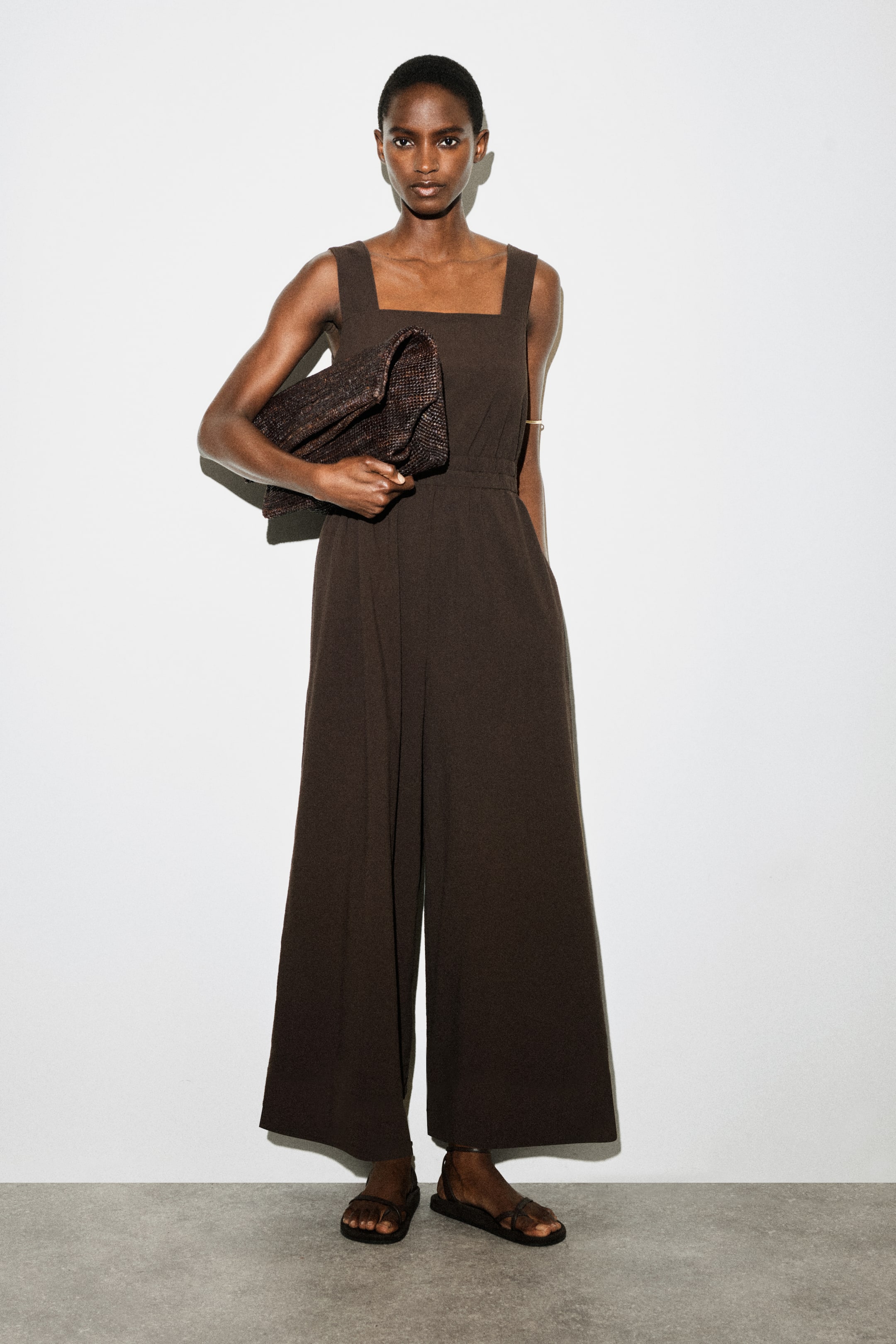 Ver imagem maior: Um macacão castanho escuro apresenta um decote quadrado, alças largas e uma cintura ajustada. As calças de perna larga fluem para baixo, e a peça é apresentada de frente.