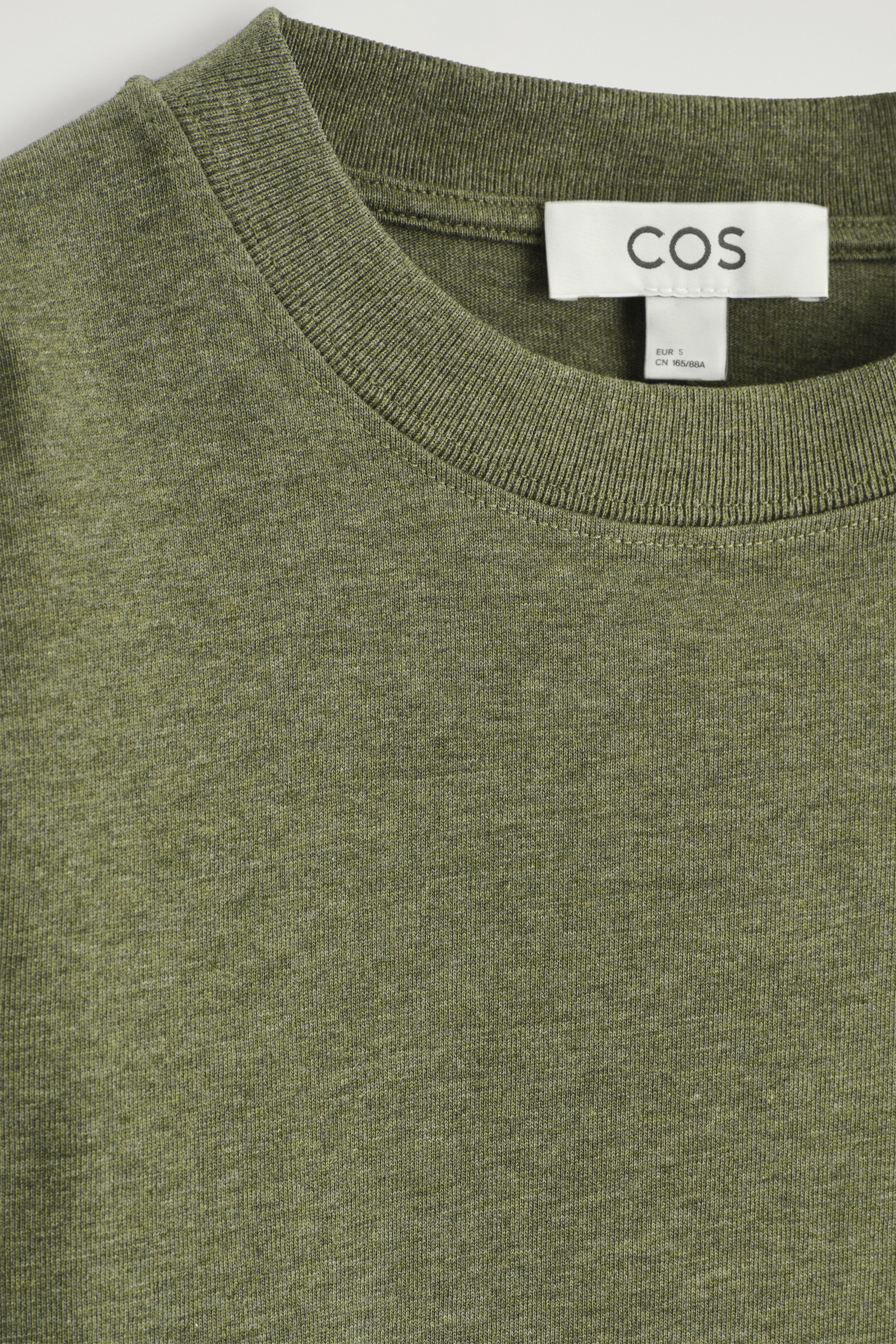 Vezi imaginea mai mare: Un tricou cu mâneci lungi de culoare verde măsliniu melanj este prezentat din FAȚĂ. Acesta are un decolteu rotund cu o margine nervurată, iar o etichetă albă de brand COS este vizibilă la spatele gâtului.