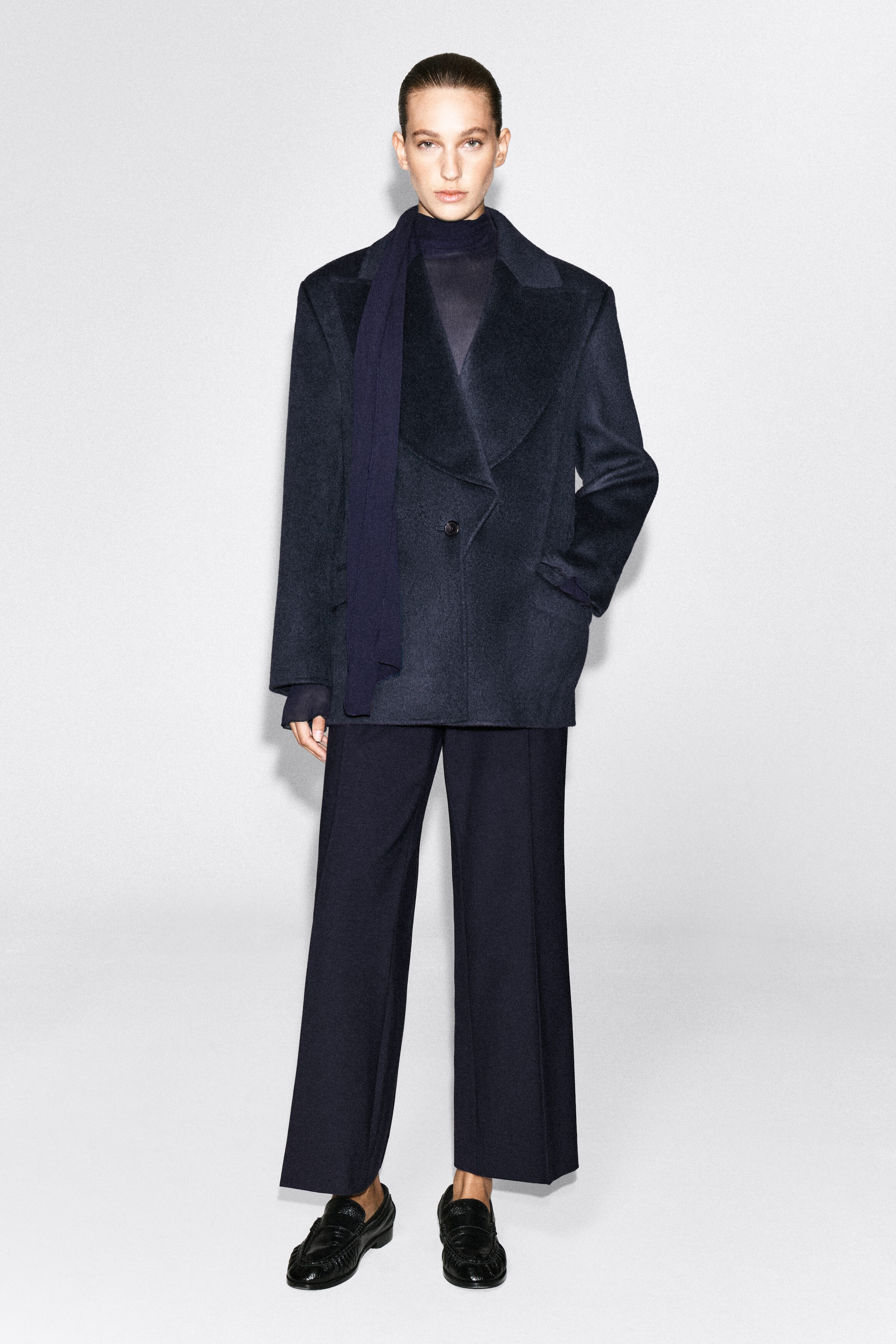 A dark navy wool-blend blazer featuring exaggerated peak lapels and a single-button fastening is being worn by the model, who is standing with one hand in her pocket. The blazer is styled with a matching draped scarf, a high-neck top, wide-leg trousers, and black loafers.