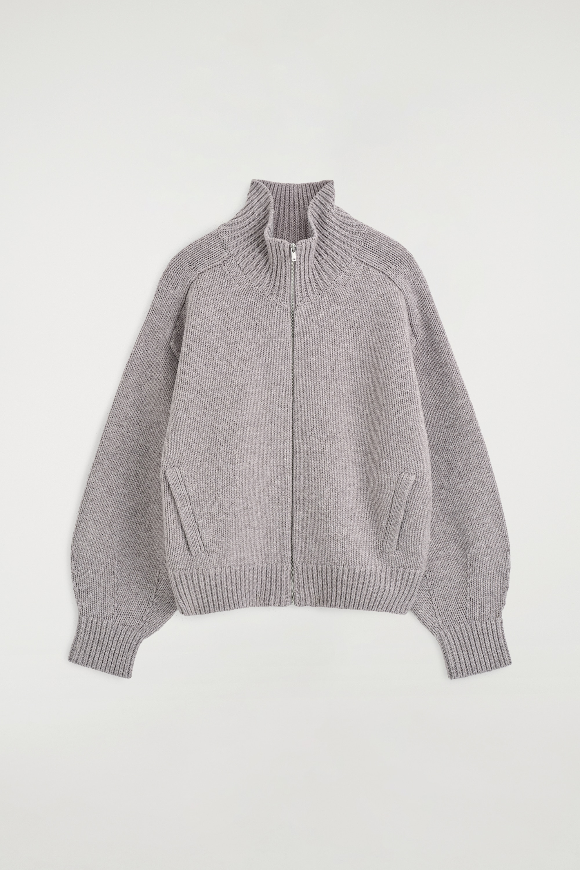 La giacca zip-up in lana con collo a imbuto arrotondato Ã¨ mostrata frontalmente, con una texture a maglia a coste grigio, una cerniera frontale argentata e un colletto alto. Ã indossata sopra un dolcevita color crema, evidenziando la vestibilitÃ  rilassata, le spalle scese e i polsini e lâorlo a costine.
