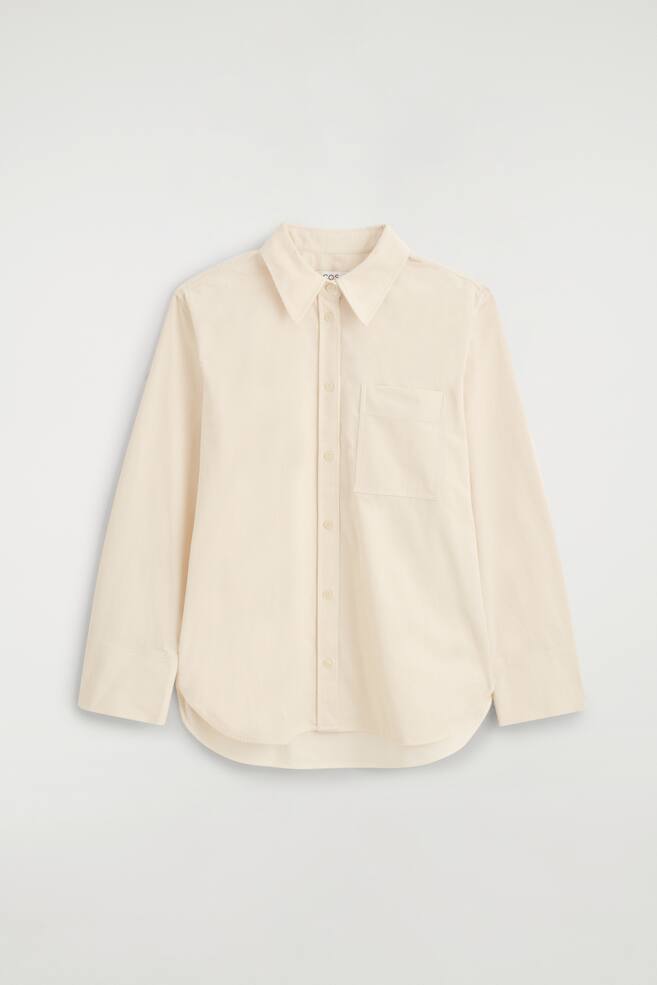Una camisa de pana de algodÃ³n color crema se muestra de frente, con cuello en punta, cierre frontal con botones y un Ãºnico bolsillo de parche en el pecho. La camisa de manga larga presenta un bajo curvado y una textura acanalada de canalÃ© fino. - 1