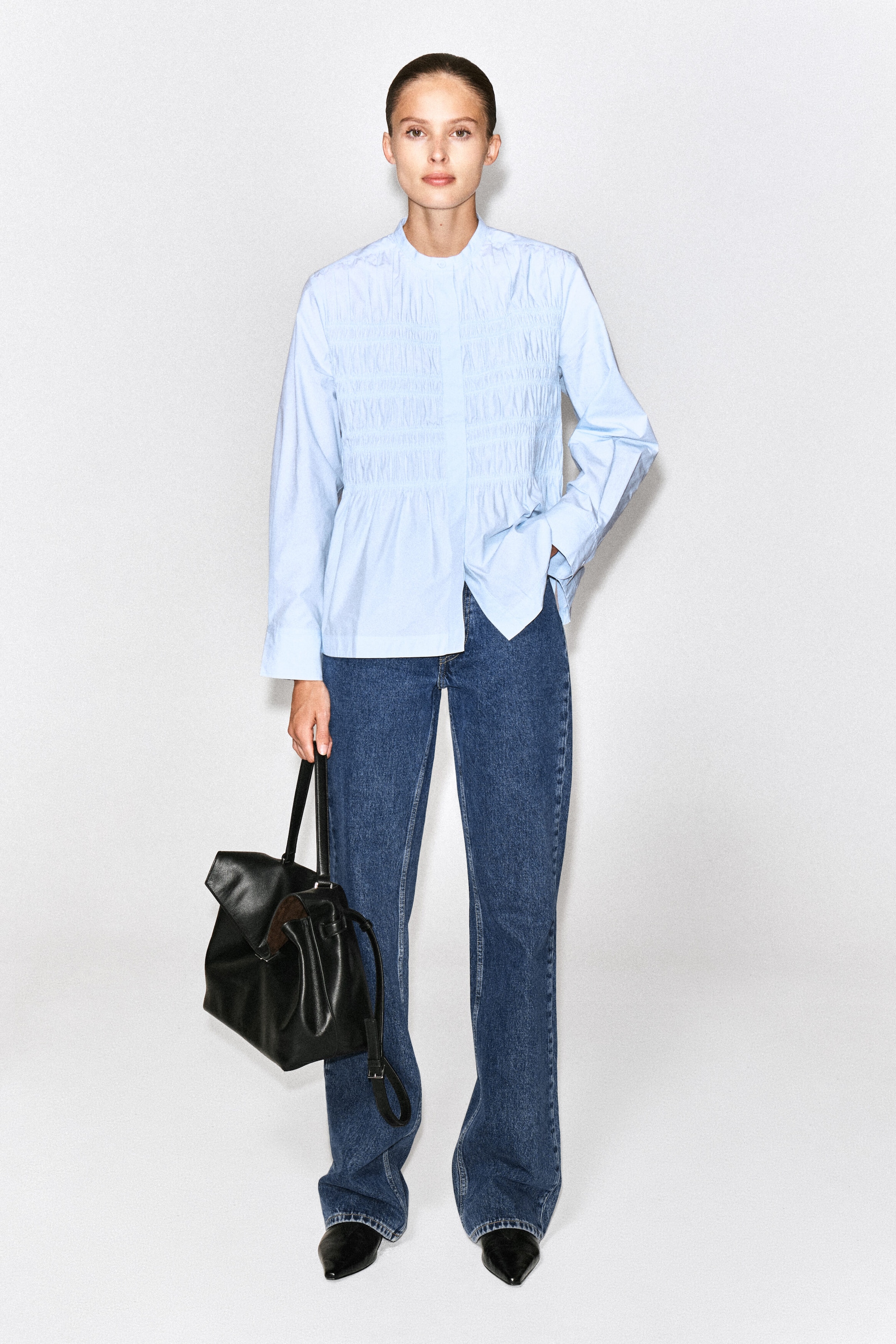 The model is wearing a light blue, long-sleeved cotton blouse featuring a band collar, horizontal ruching across the bodice, and a flared hem. The blouse is styled with dark blue jeans, and the model is carrying a black leather handbag. The model is wearing a light blue, long-sleeved cotton blouse featuring a band collar, horizontal ruching across the bodice, and a flared hem. The blouse is styled with dark blue jeans, and the model is carrying a black leather handbag.