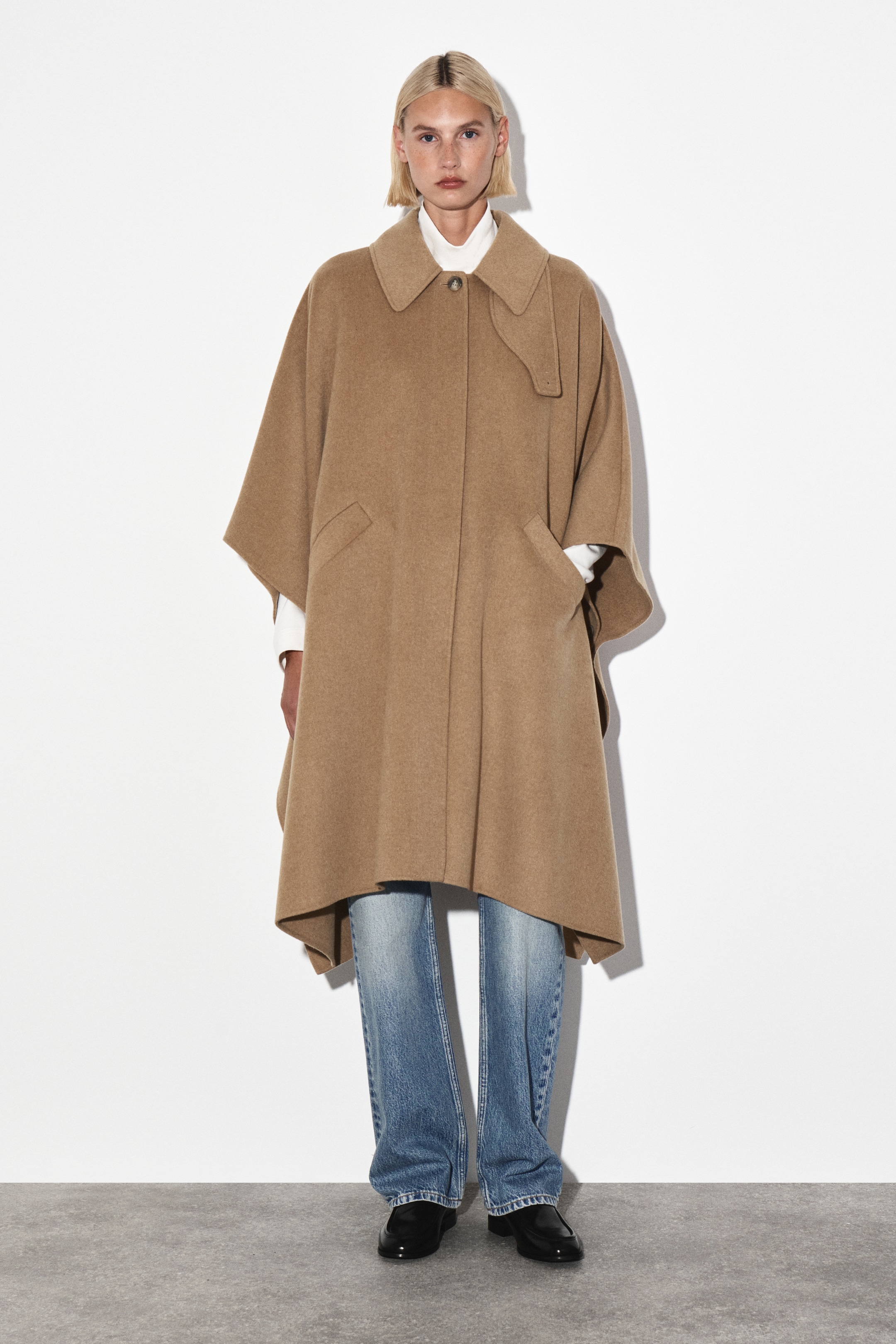 Agrandir l'image: Une cape en chameau est orientée vers l'avant, avec un col pointu doté d'un bouton supérieur visible et d'une patte décorative boutonnée sur le côté droit. Elle est conçue avec deux poches plaquées avant inclinées et un ourlet asymétrique, tombant jusqu'aux genoux, avec une chemise à col blanc visible en dessous.