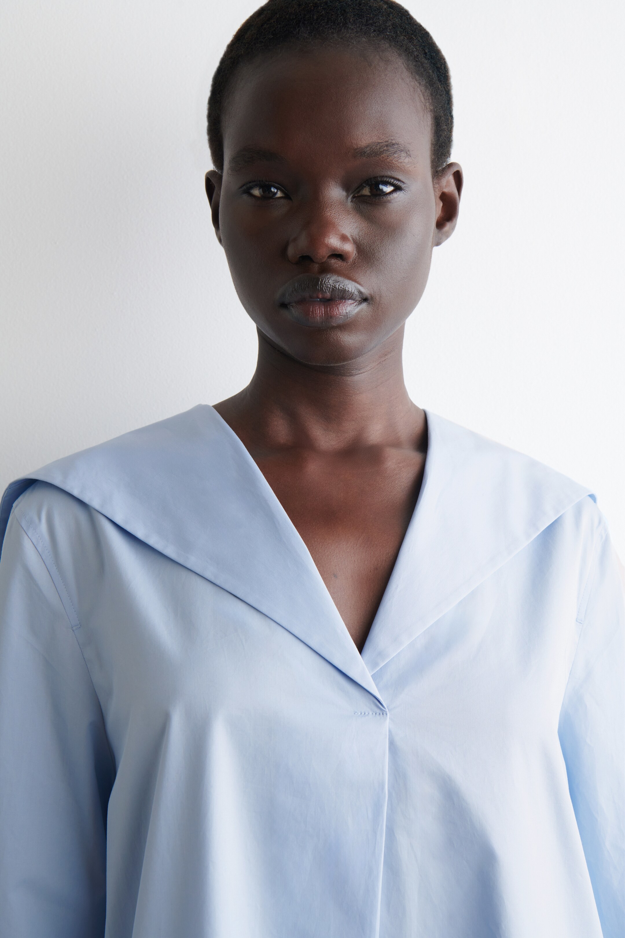 Agrandir l'image: Une chemise bleu clair à manches longues est portée, vue de face. Elle présente un col large et plat qui forme un profond décolleté en V, et le tissu net montre des plis subtils sur la poitrine.
