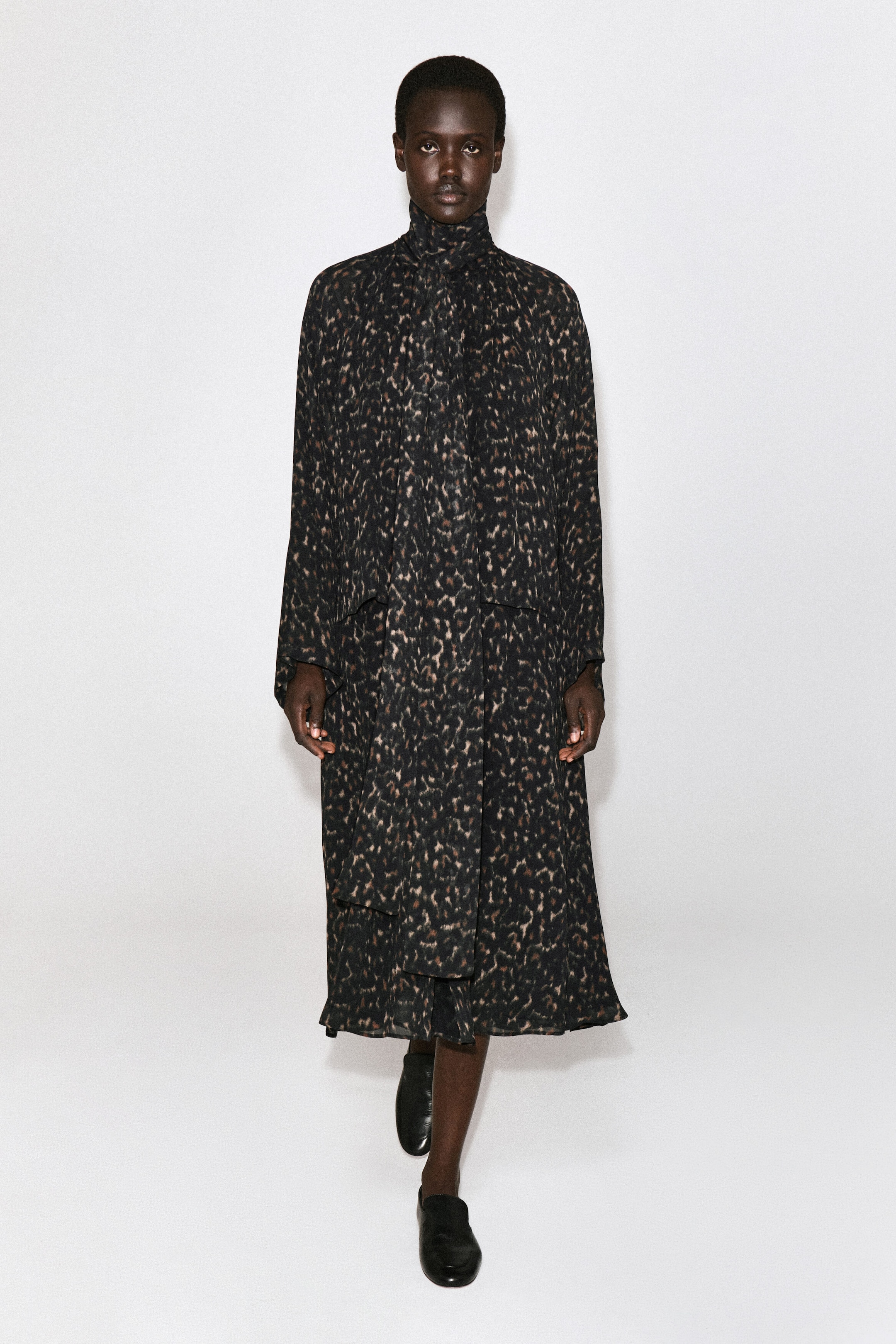 The leopard-print fluid dress with scarf detail is shown from the front, featuring a high neckline with an integrated draped scarf detail, long sleeves, and a loose-fitting midi-length silhouette. The dress is styled with black leather slip-on shoes. The leopard-print fluid dress with scarf detail is shown from the front, featuring a high neckline with an integrated draped scarf detail, long sleeves, and a loose-fitting midi-length silhouette. The dress is styled with black leather slip-on shoes.
