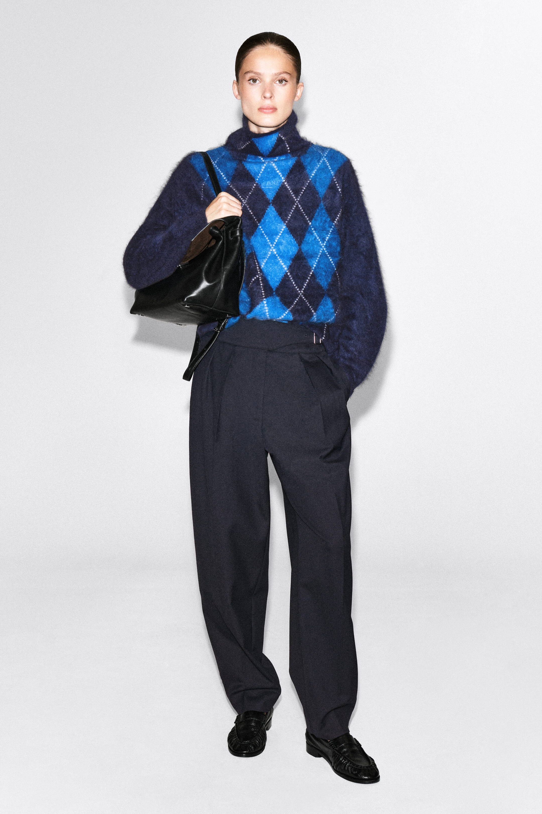 A brushed cashmere funnel-neck jumper featuring a blue and navy argyle pattern is being worn by the model, tucked into dark, wide-leg trousers. The model is carrying a black leather shoulder bag that rests against the side of the jumper. A brushed cashmere funnel-neck jumper featuring a blue and navy argyle pattern is being worn by the model, tucked into dark, wide-leg trousers. The model is carrying a black leather shoulder bag that rests against the side of the jumper.