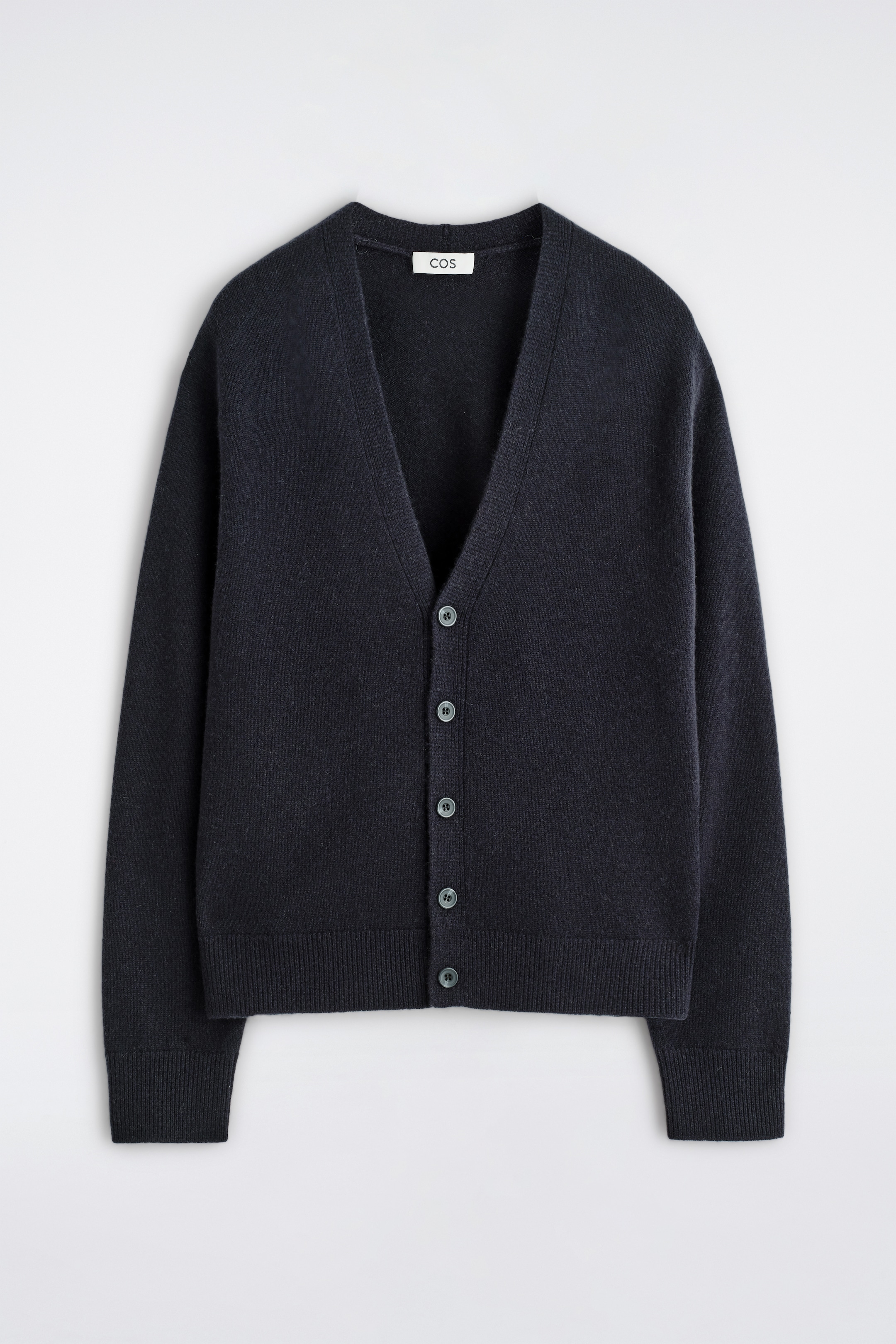 Größeres Bild ansehen: Eine navyfarbene Kaschmirstrickjacke wird vorne präsentiert, mit einem V-Ausschnitt und einer Knopfleiste mit sieben dunklen, runden Knöpfen. Sie zeigt eine fein gestrickte Textur, mit gerippten Bündchen und einem gerippten Saum, und ein kleines weißes 'COS'-Etikett ist am hinteren Ausschnitt sichtbar.