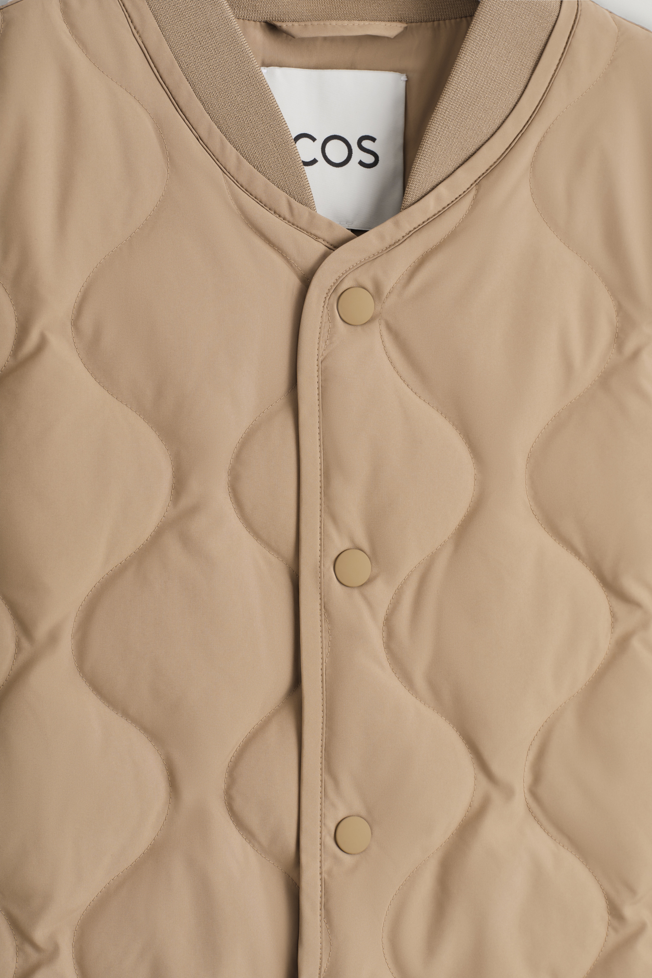 Agrandir l'image: Un gilet beige est présenté de face, avec un motif matelassé ondulé. Il a un col côtelé et une patte de boutonnage centrale à l'avant, fermée par trois boutons-pression assortis.