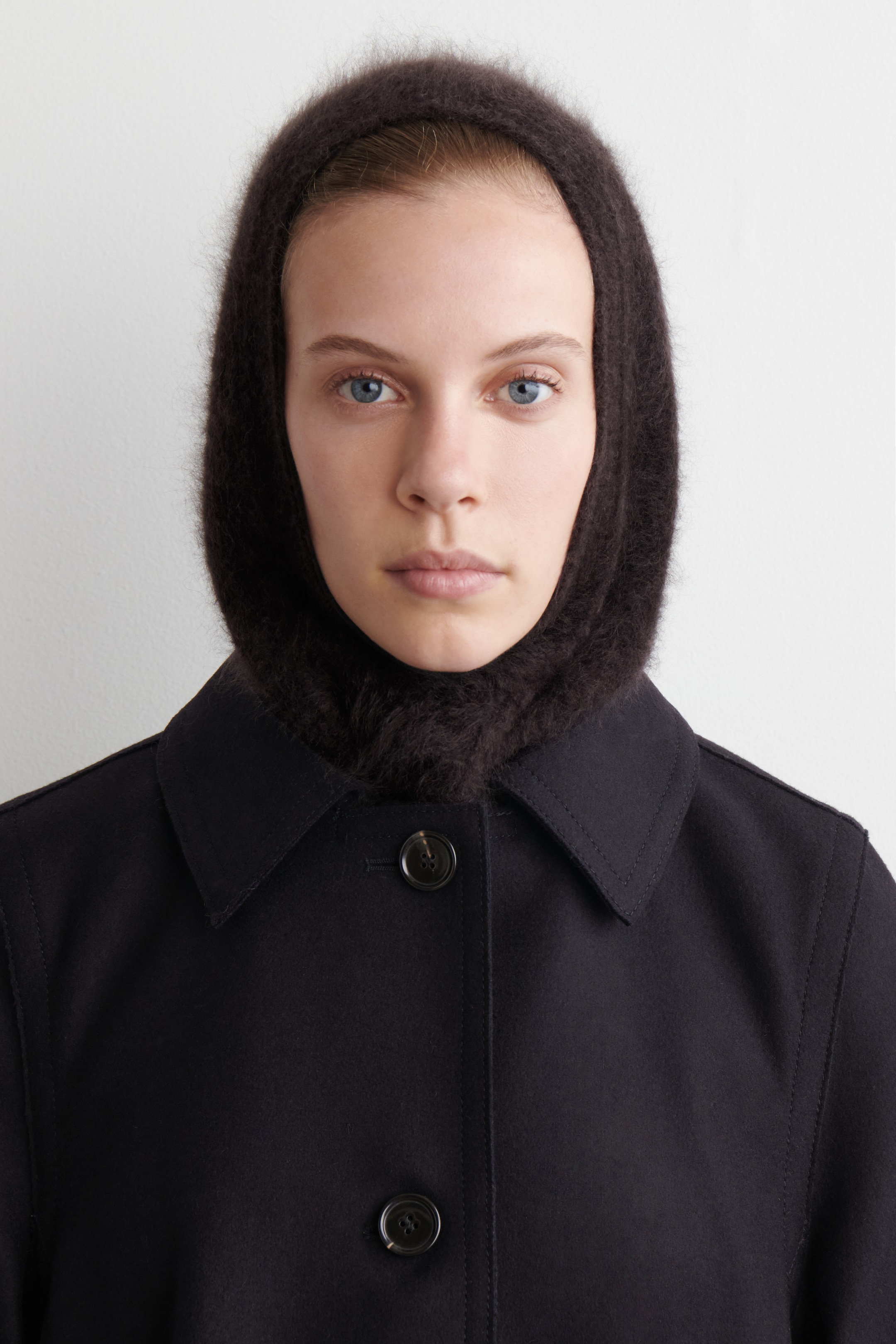 Agrandir l'image: Une cagoule en tricot brun foncé à longs poils encadre le visage et couvre la tête et le cou. Elle est portée sous un manteau noir, qui présente un col classique et deux boutons sombres et visibles sur le devant.