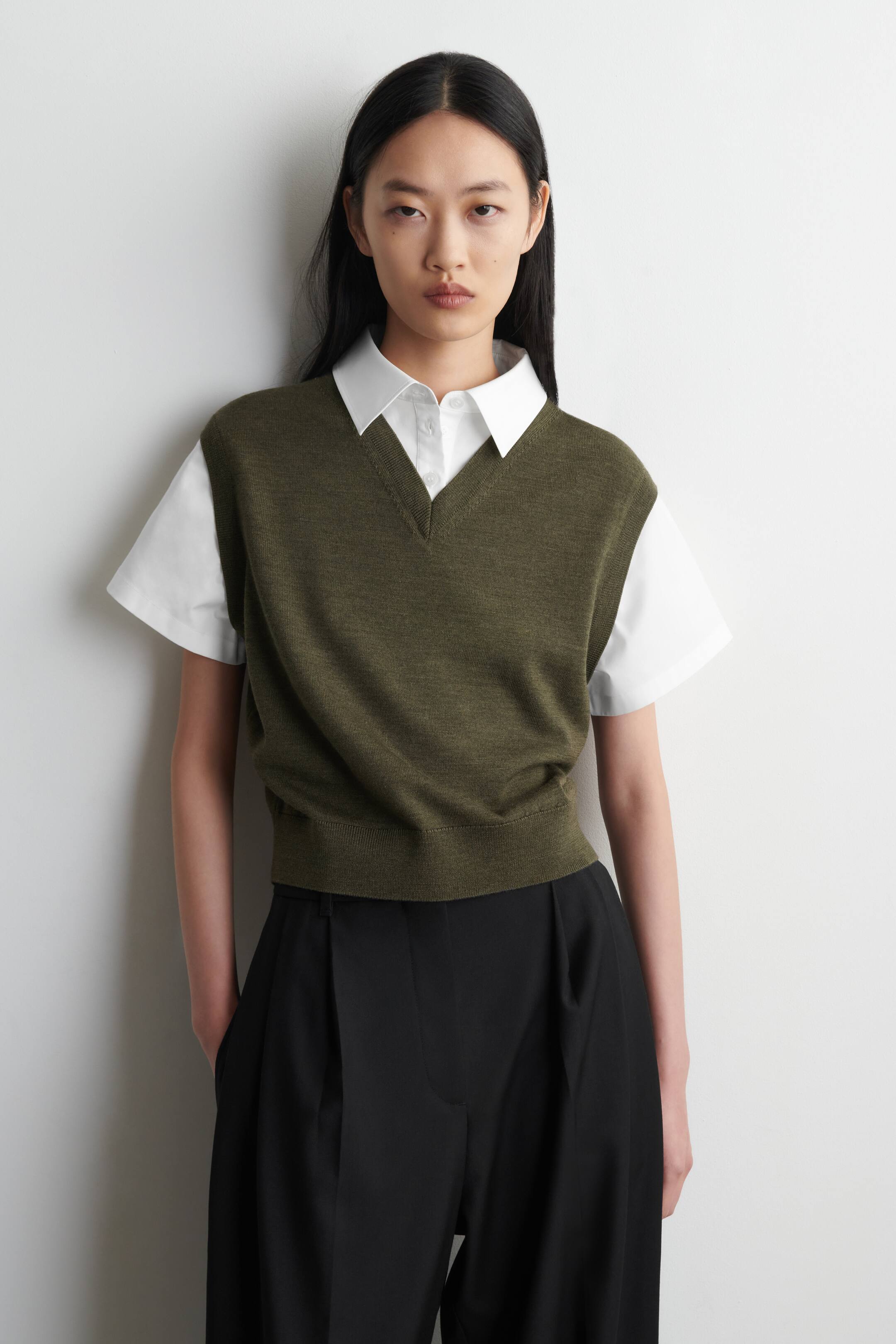 הצגת תמונה גדולה יותר: אפוד סוודר בצבע ירוק זית עם מחשוף V, בעל מרקם סריגה עדין, מחשוף בצורת V ושוליים מצולעים, מוצג מהחזית ומונח על חולצה לבנה עם שרוולים קצרים וצווארון.