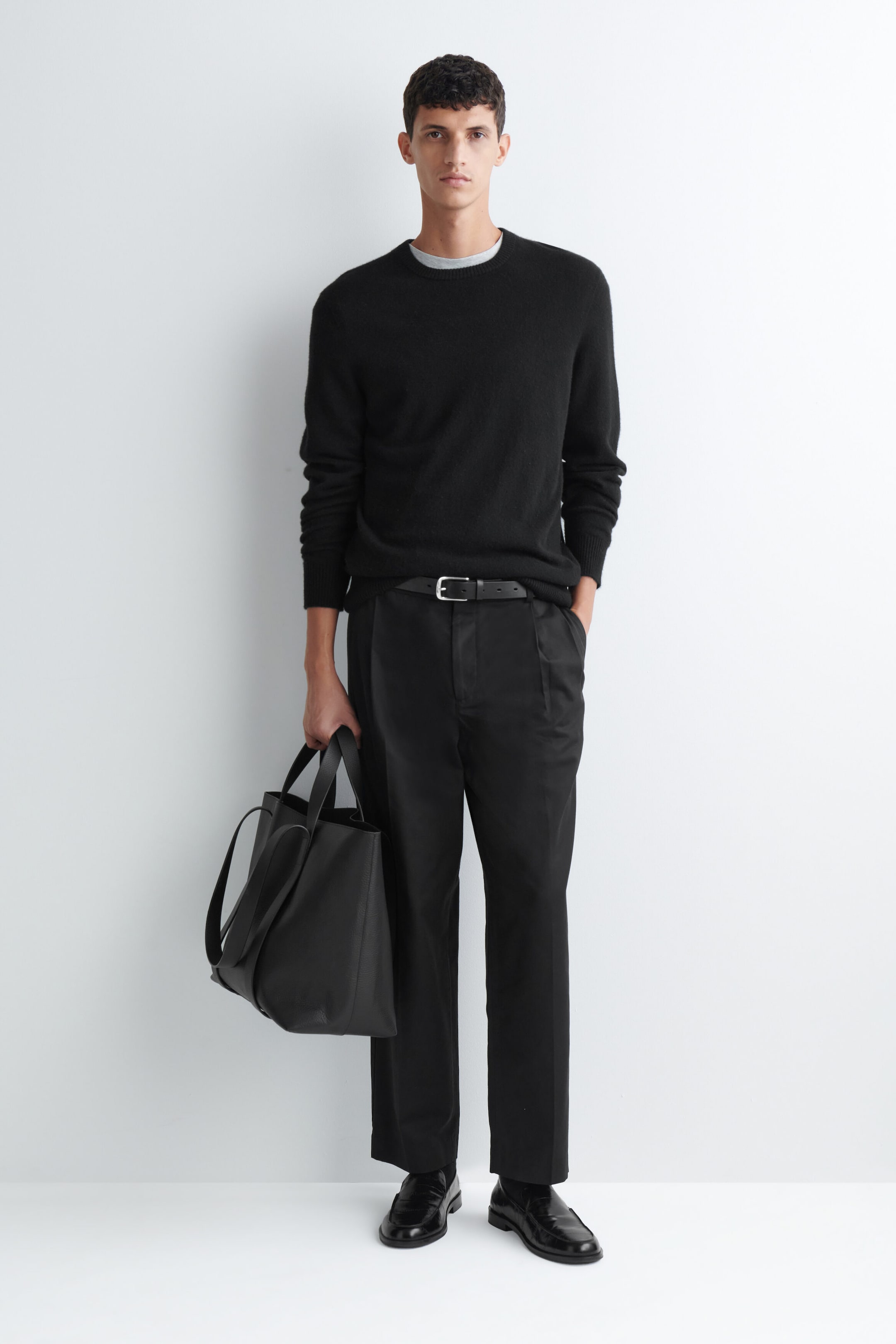 Ein schwarzer Pullover mit Rundhalsausschnitt und langen Ärmeln wird vom Model getragen und mit einer schwarzen, maßgeschneiderten Hose sowie einem schwarzen Ledergürtel kombiniert. Das Model hält außerdem eine schwarze Leder-Tote-Bag an den oberen Henkeln und rundet damit den monochromen Look ab. Ein schwarzer Pullover mit Rundhalsausschnitt und langen Ärmeln wird vom Model getragen und mit einer schwarzen, maßgeschneiderten Hose sowie einem schwarzen Ledergürtel kombiniert. Das Model hält außerdem eine schwarze Leder-Tote-Bag an den oberen Henkeln und rundet damit den monochromen Look ab.