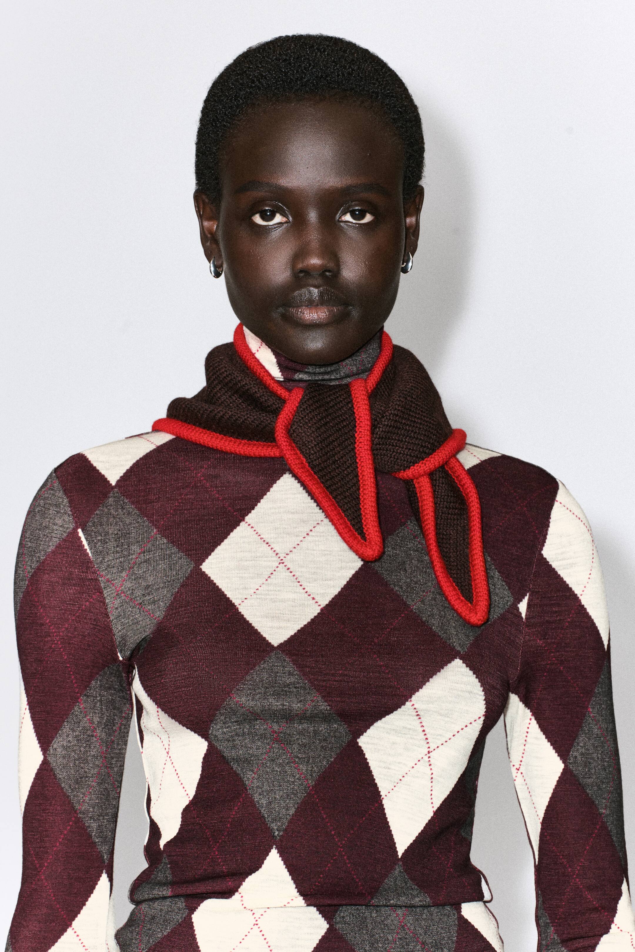Grotere afbeelding bekijken: De driehoekige sjaal van merinowol met contrasterende randen sluit nauw rond de hals in vooraanzicht, gestyled over een coltrui met argylemotief. De donkerbruine gebreide sjaal toont een levendige rode contrasterende boord langs de randen, terwijl de geknoopte uiteinden over de borst vallen.