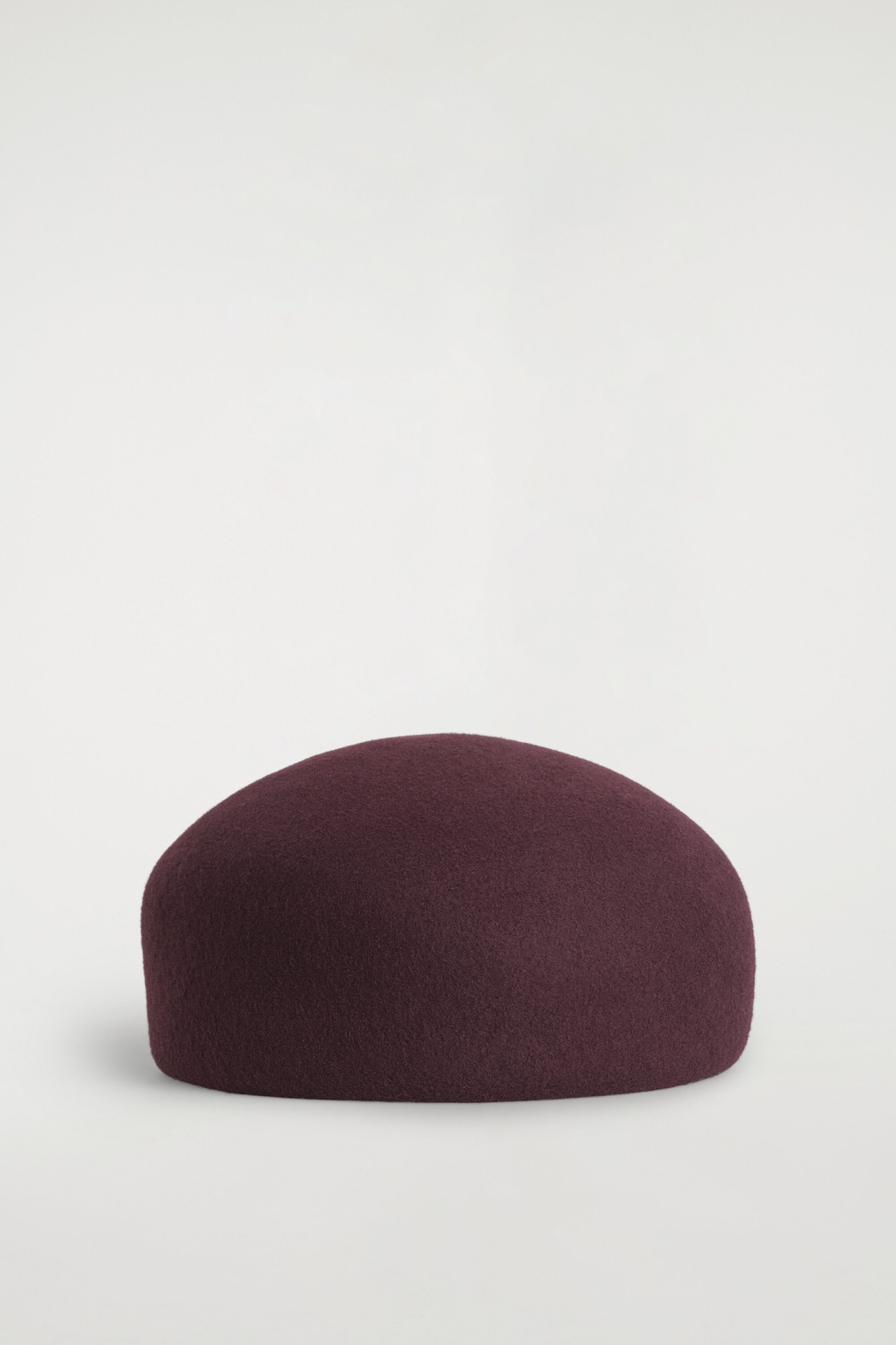A structured, dome-shaped beret in deep burgundy boiled wool is lying flat, showcasing its rounded, sculptural silhouette from the front.
