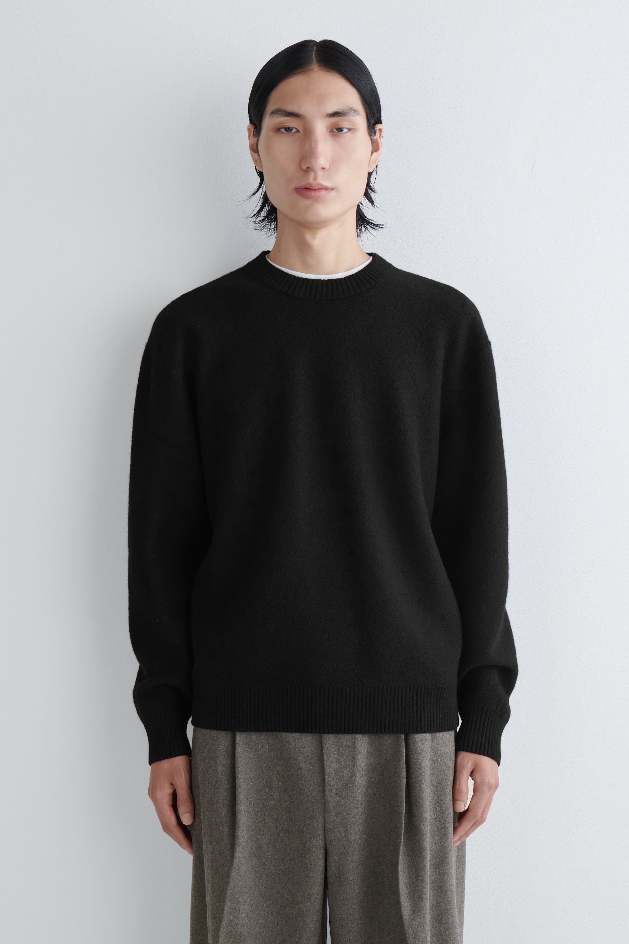 Grösseres Bild ansehen: Ein schwarzer Pullover mit Rundhalsausschnitt wird in der Vorderansicht präsentiert und zeichnet sich durch ein subtil strukturiertes Strickmuster aus. Er ist mit einem gerippten Rundhalsausschnitt, gerippten Bündchen und einem gerippten Saum gestaltet und wird über einem weißen T-Shirt getragen, dessen Kragen sichtbar ist.