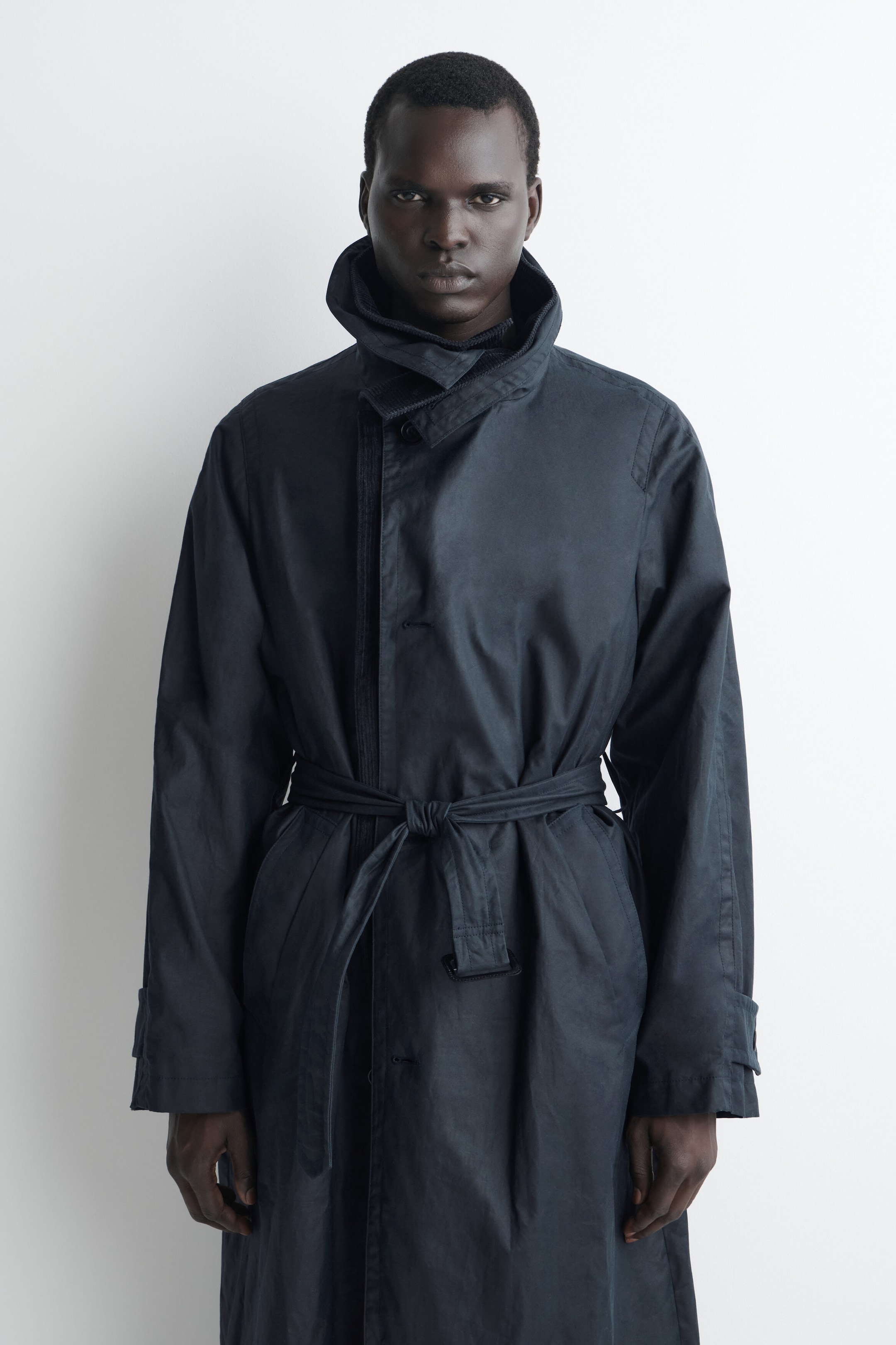 A midnight blue waxed cotton trench coat is shown from the front, featuring a high funnel-neck collar and a matching self-tie belt at the waist. The relaxed-fit coat is styled closed, highlighting its textured fabric and concealed placket. A midnight blue waxed cotton trench coat is shown from the front, featuring a high funnel-neck collar and a matching self-tie belt at the waist. The relaxed-fit coat is styled closed, highlighting its textured fabric and concealed placket.