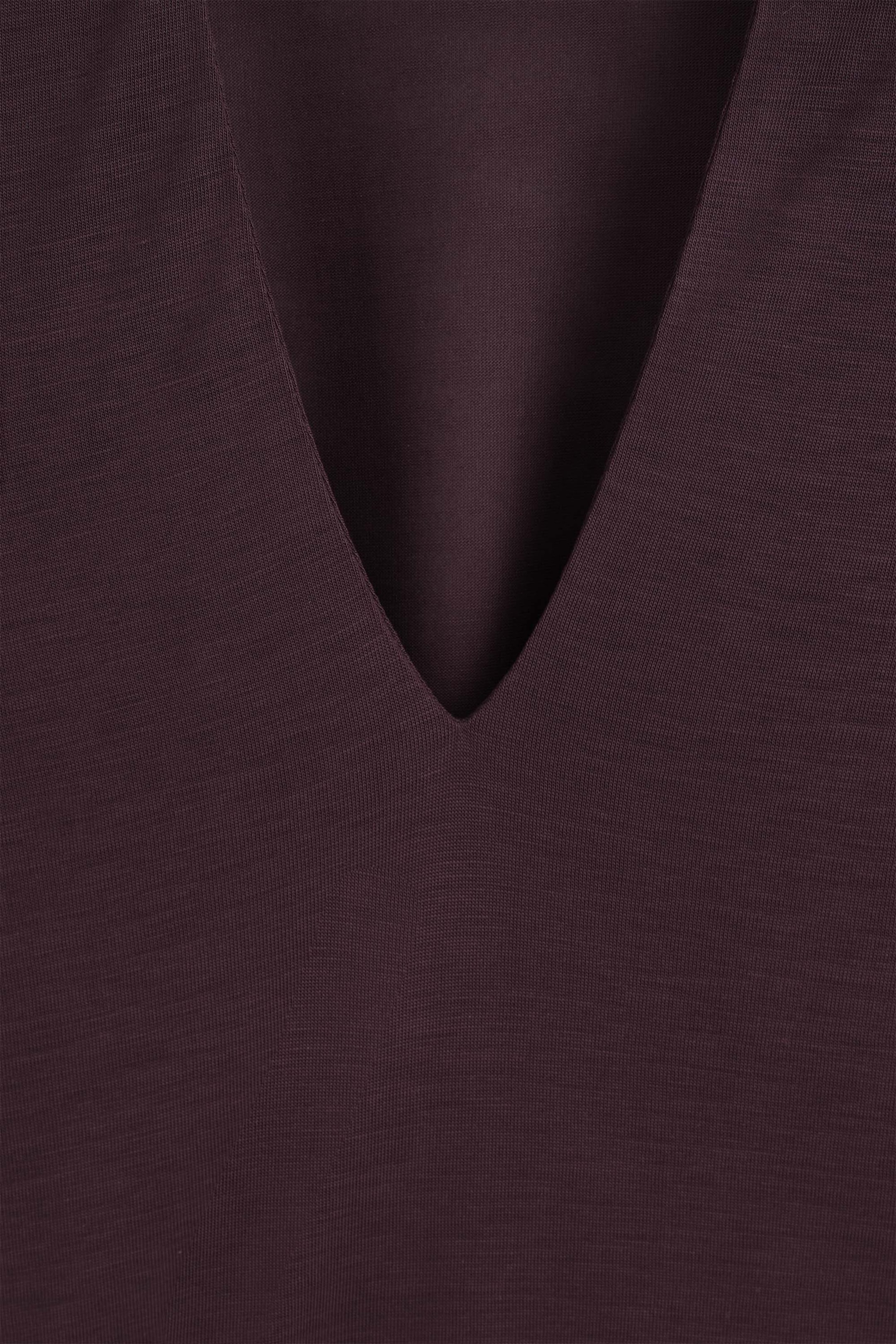 A close-up of a dark plum-colored jersey top showcasing the clean-edged V-neckline, with a draped layer of matching fabric visible behind the opening to highlight its smooth, fine-knit texture. A close-up of a dark plum-colored jersey top showcasing the clean-edged V-neckline, with a draped layer of matching fabric visible behind the opening to highlight its smooth, fine-knit texture.