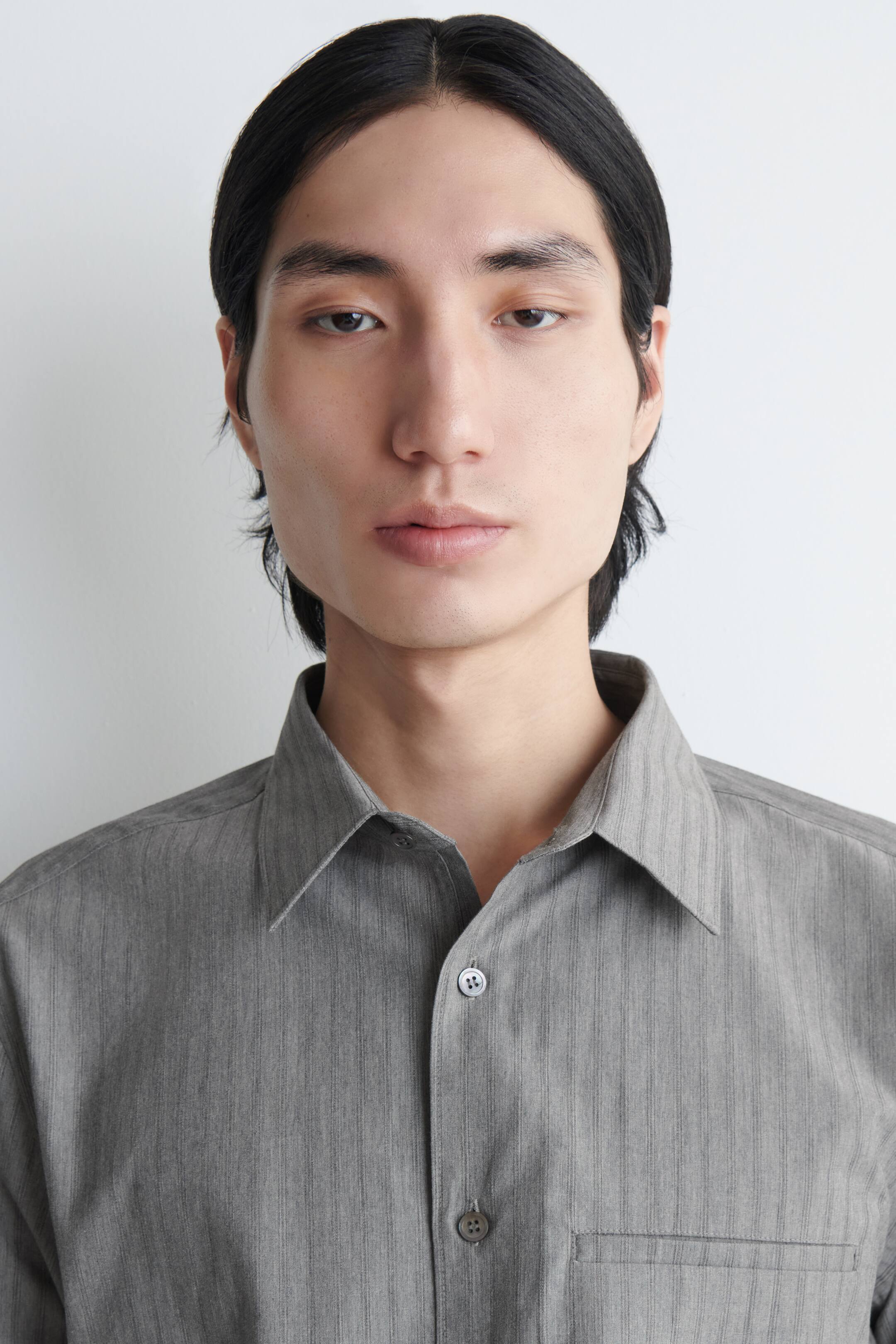 View larger image: A grey button-up shirt with a subtle vertical texture is shown front-on, featuring a spread collar with the top button undone, grey buttons down the front placket, and a patch pocket on the left chest.