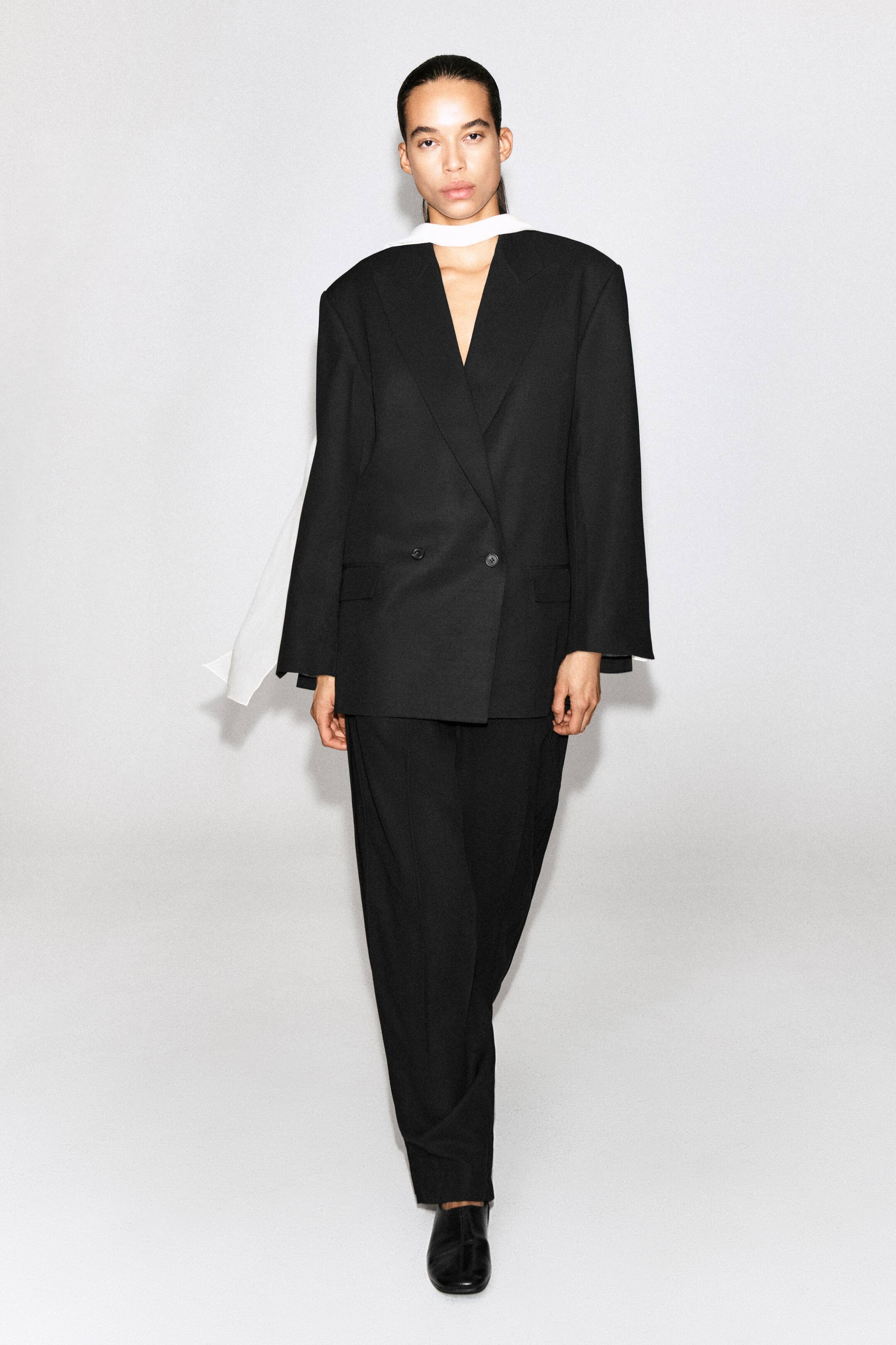 Grotere afbeelding bekijken: Deze zwarte, dubbelrijige blazer met oversized silhouet, gestructureerde schouders en een sluiting met twee knopen aan de voorkant wordt getoond vanuit een vooraanzicht. De blazer wordt gestyled met contrasterende witte stof die rond de hals is gedrapeerd en gecombineerd met een bijpassende zwarte broek.