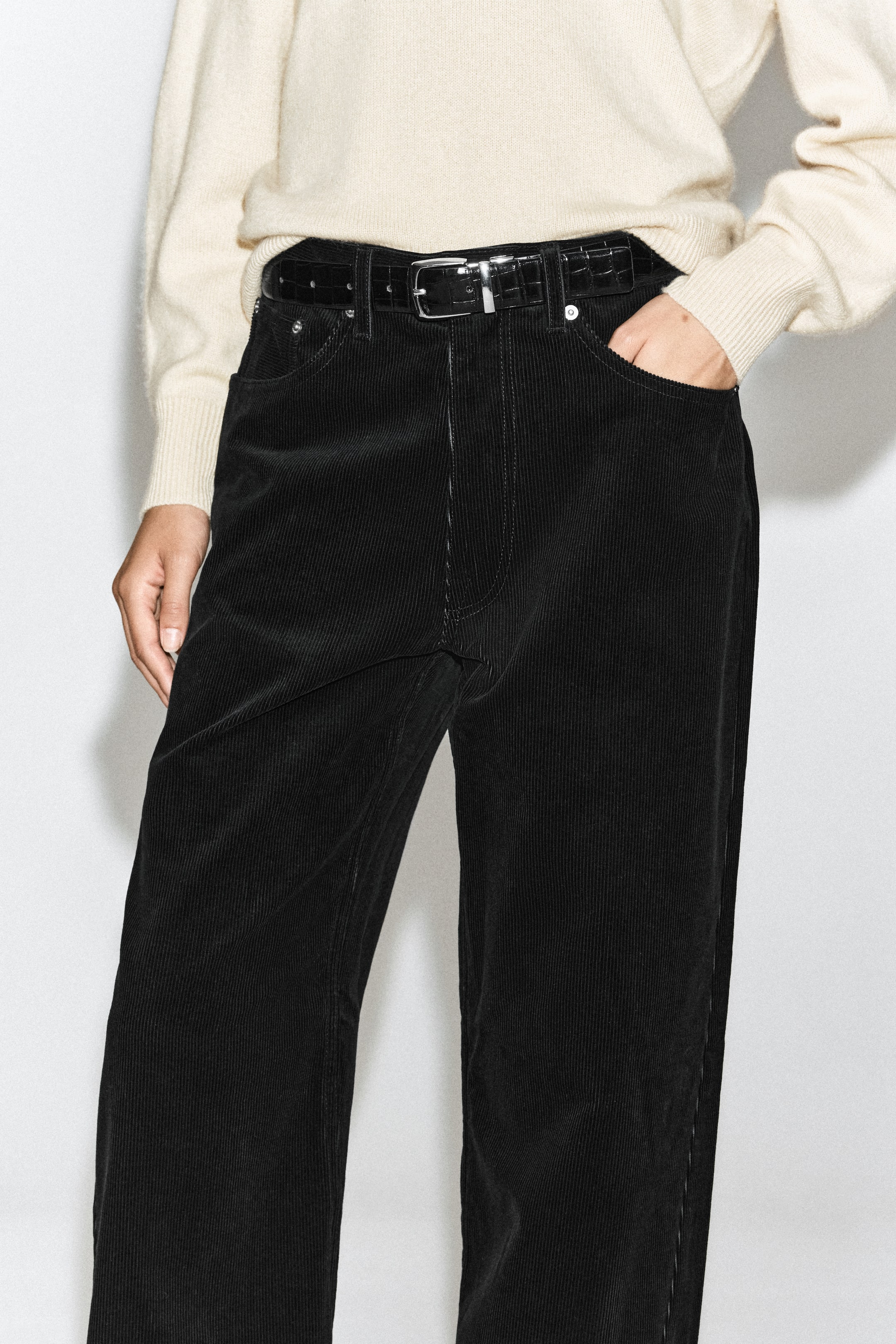 Visualizza immagine più grande: I jeans Facade in velluto a coste dritti, neri, sono indossati dalla modella con un maglione color crema infilato nei pantaloni e una cintura nera texturizzata. I jeans presentano una texture a coste in velluto, passanti per cintura e tasche frontali classiche, con la modella che appoggia una mano in una tasca.