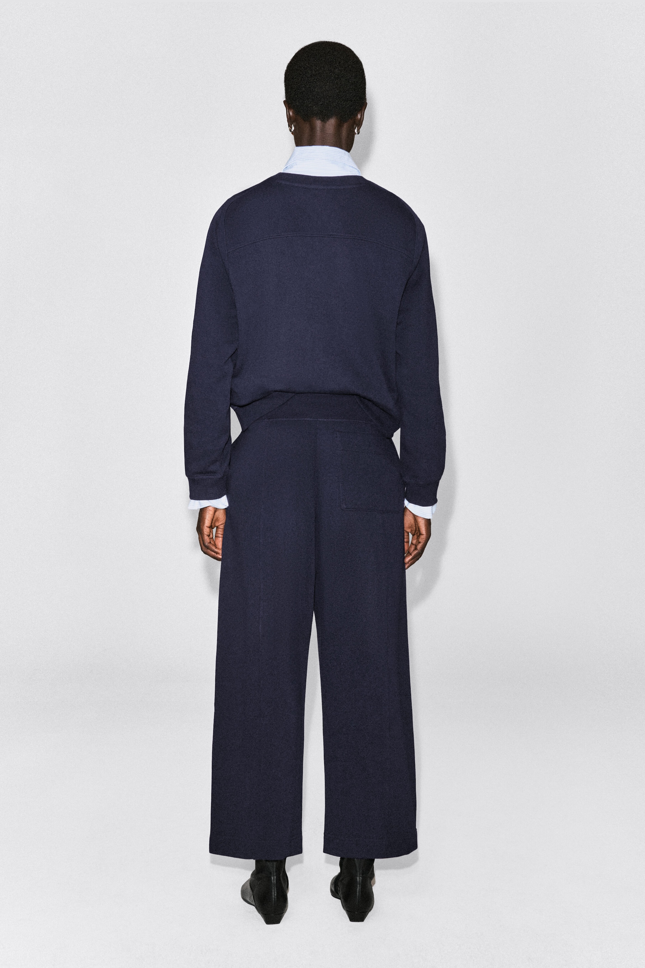 A navy blue cotton sweatshirt is shown from the back, featuring a horizontal seam across the upper back, ribbed cuffs, and a ribbed hem. The sweatshirt is layered over a light blue collared shirt and paired with matching dark blue wide-leg trousers. A navy blue cotton sweatshirt is shown from the back, featuring a horizontal seam across the upper back, ribbed cuffs, and a ribbed hem. The sweatshirt is layered over a light blue collared shirt and paired with matching dark blue wide-leg trousers.