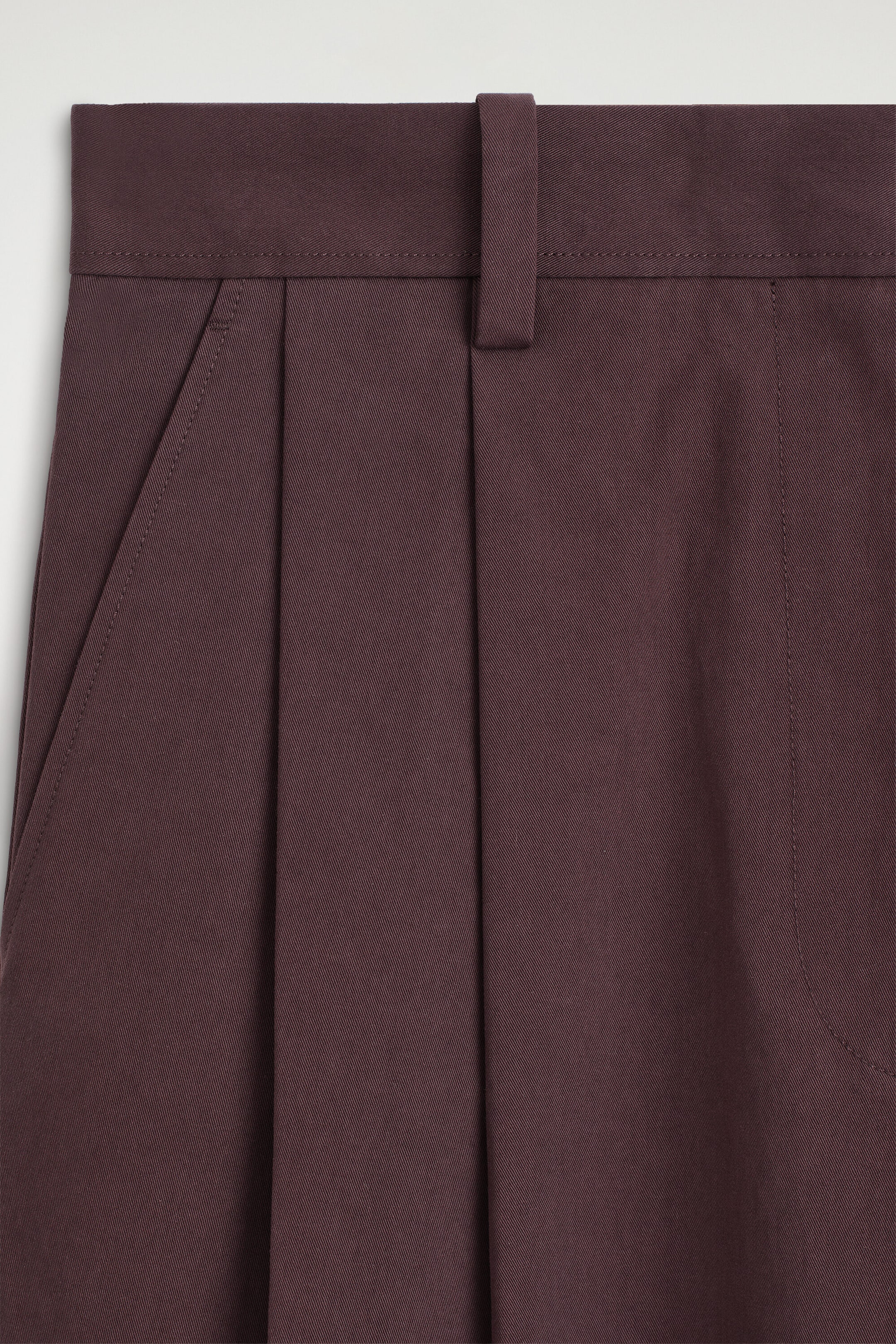 Cotton cropped barrel-leg pants in a deep plum shade are shown in a close-up front view of the waistband, featuring a center belt loop, deep front pleats, and a slanted side pocket. The structured woven fabric shows a subtle diagonal twill texture. Cotton cropped barrel-leg pants in a deep plum shade are shown in a close-up front view of the waistband, featuring a center belt loop, deep front pleats, and a slanted side pocket. The structured woven fabric shows a subtle diagonal twill texture.