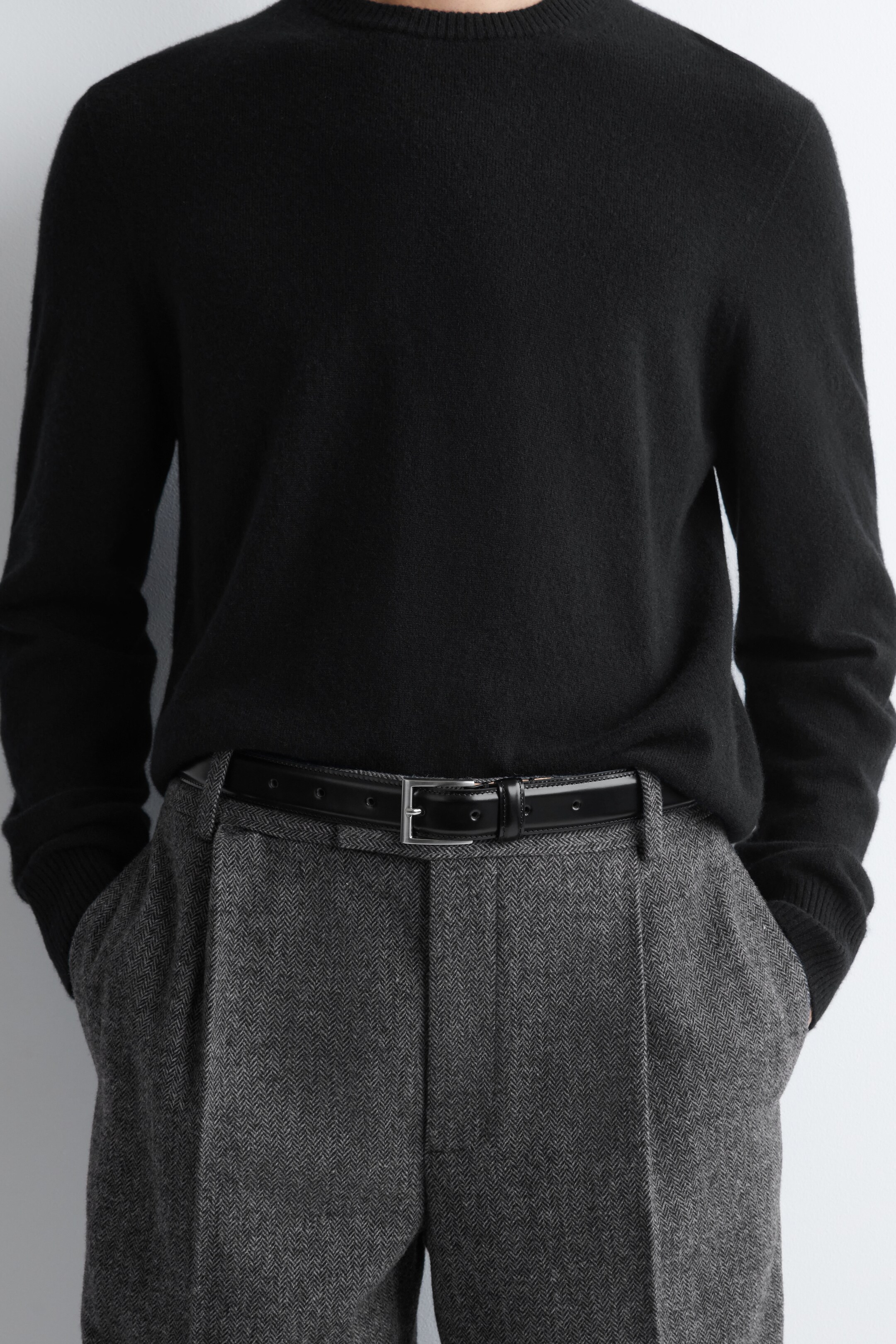 Agrandir l'image: Un pull noir à col rond est porté, rentré dans un pantalon gris en chevron. Le pantalon présente des plis à l'avant, et une ceinture noire avec une boucle rectangulaire argentée le maintient à la taille.