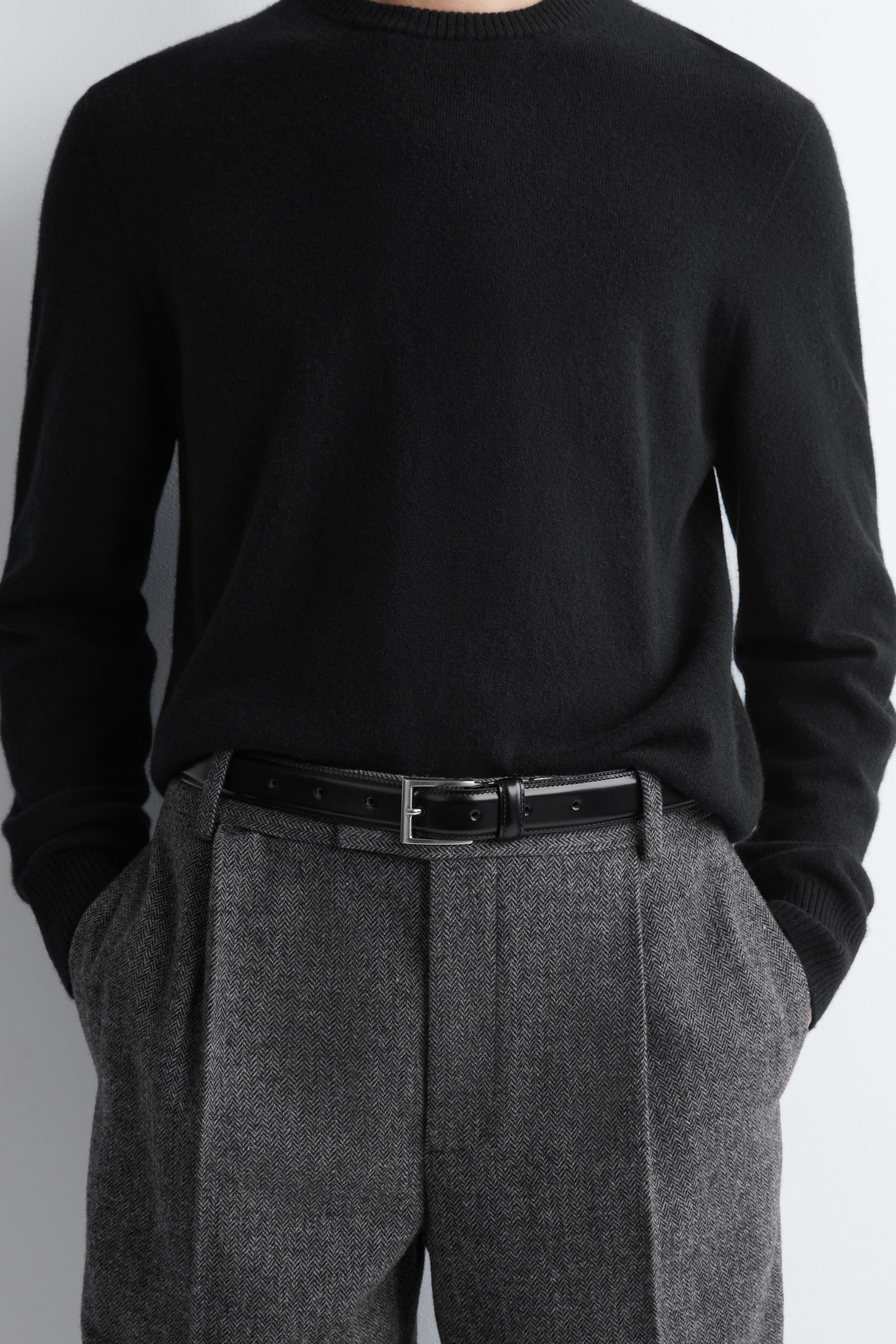 View larger image: A black crew-neck sweater is being worn, tucked into gray herringbone trousers. The trousers are featuring front pleats, and a black belt with a rectangular silver-toned buckle is securing them at the waist.