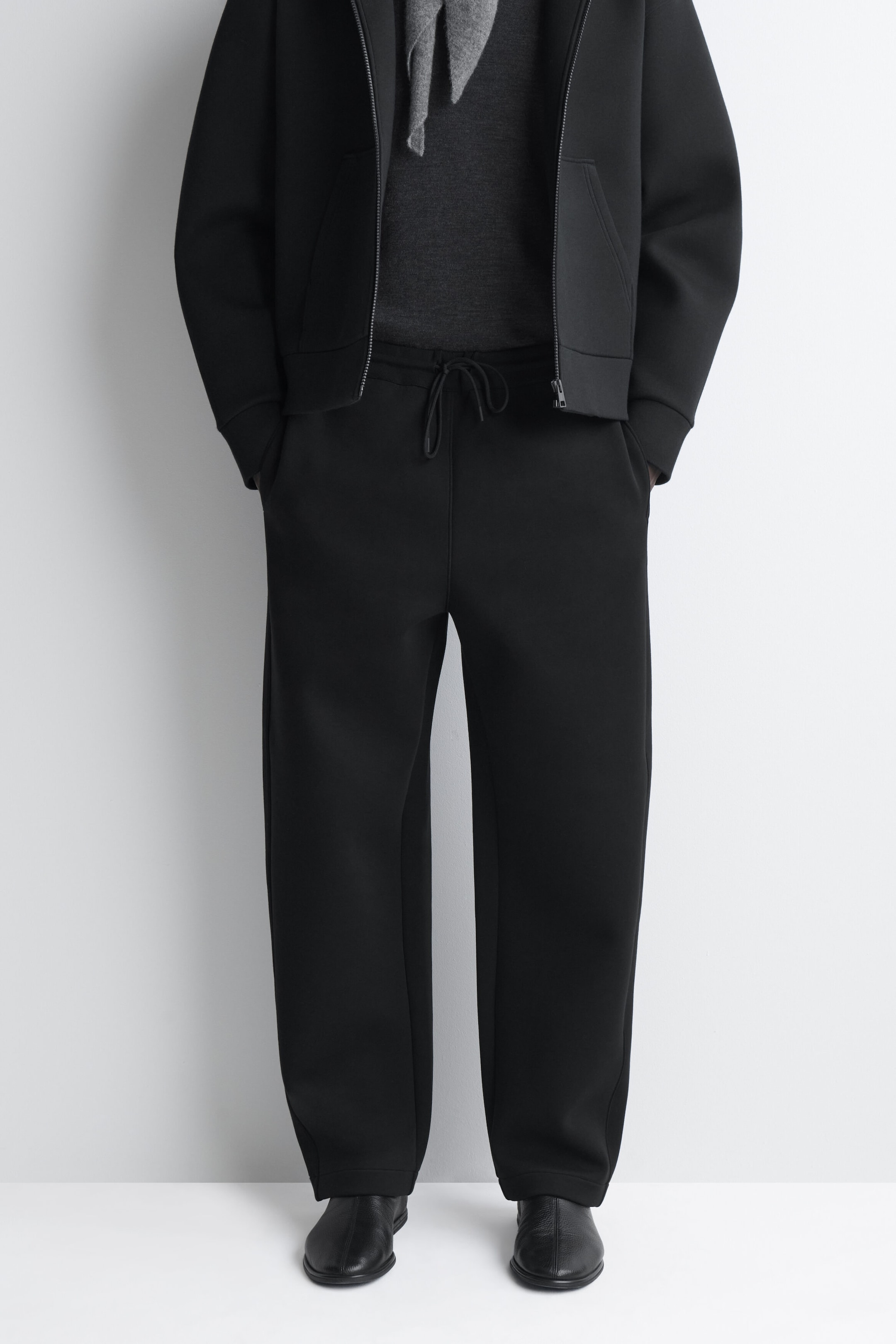 Black structured sweatpants feature a relaxed, straight-leg fit and an elasticated waistband with a front drawstring tie. Shown from the waist down to the hem, the pants are styled with a matching black zip-up jacket and black leather shoes to highlight their clean, modern drape.