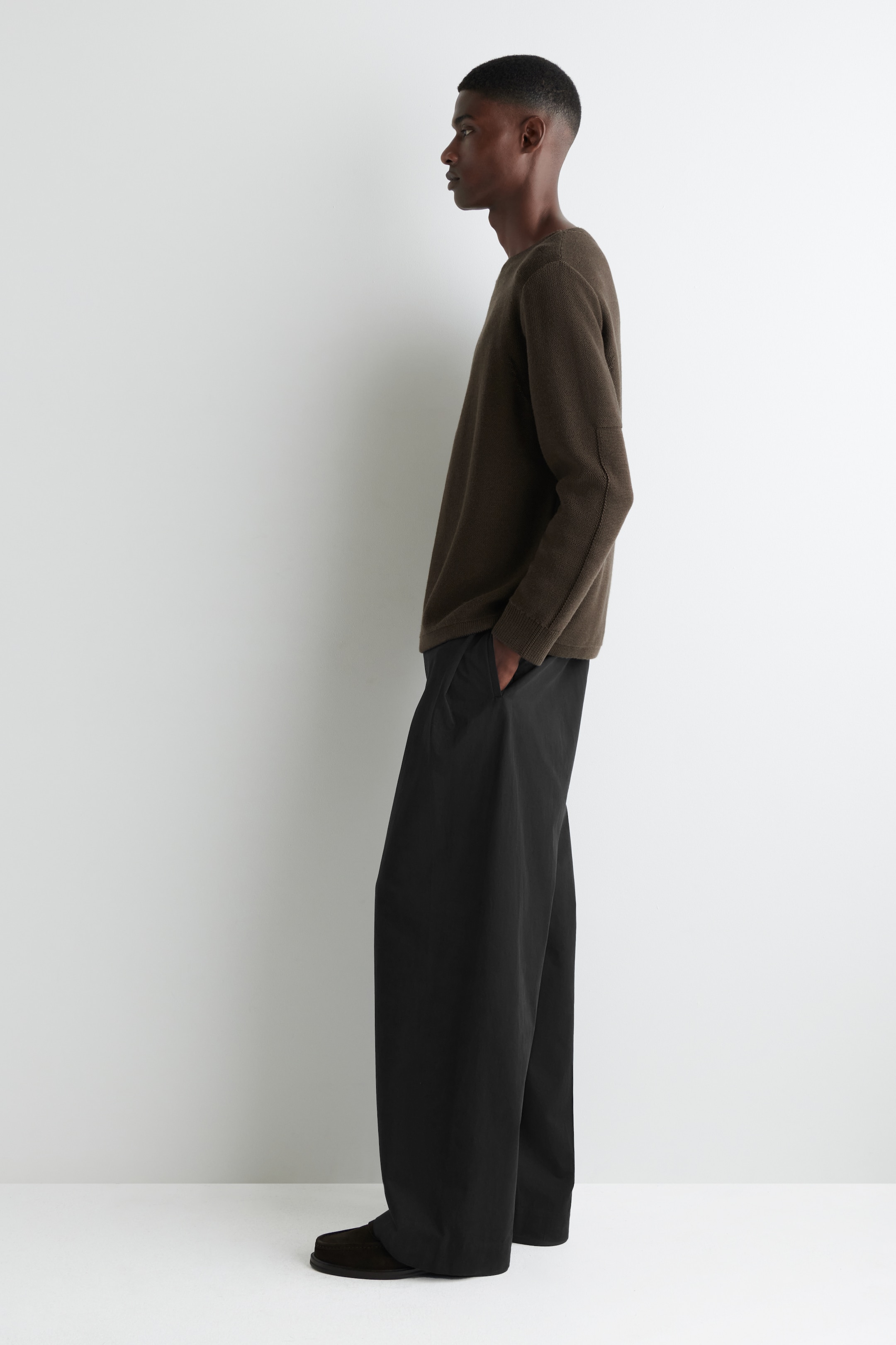 Grösseres Bild ansehen: Ein dunkelbrauner langärmeliger Pullover wird mit schwarzen Hosen mit weitem Bein präsentiert, von der linken Seite betrachtet. Der Pullover hat eine sichtbare Naht, die den Arm hinunterläuft, und die Hosen zeigen eine lockere, fließende Passform.