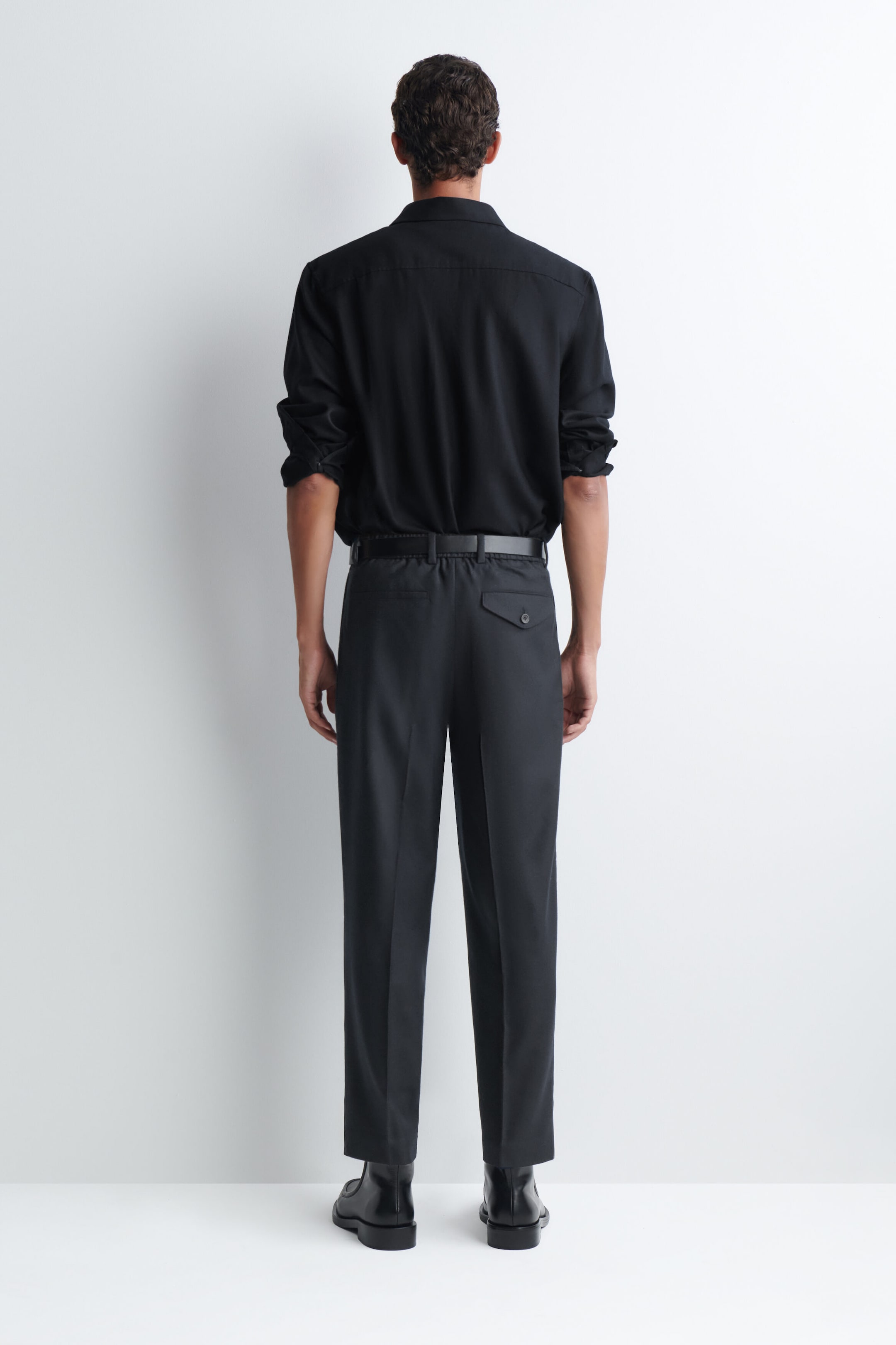 The model is wearing dark gray tailored trousers, shown from the back, styled with a tucked-in black long-sleeve shirt, a black belt, and black boots. The trousers feature a straight-leg cut, pressed leg creases, and a single back pocket with a buttoned flap. The model is wearing dark gray tailored trousers, shown from the back, styled with a tucked-in black long-sleeve shirt, a black belt, and black boots. The trousers feature a straight-leg cut, pressed leg creases, and a single back pocket with a buttoned flap.