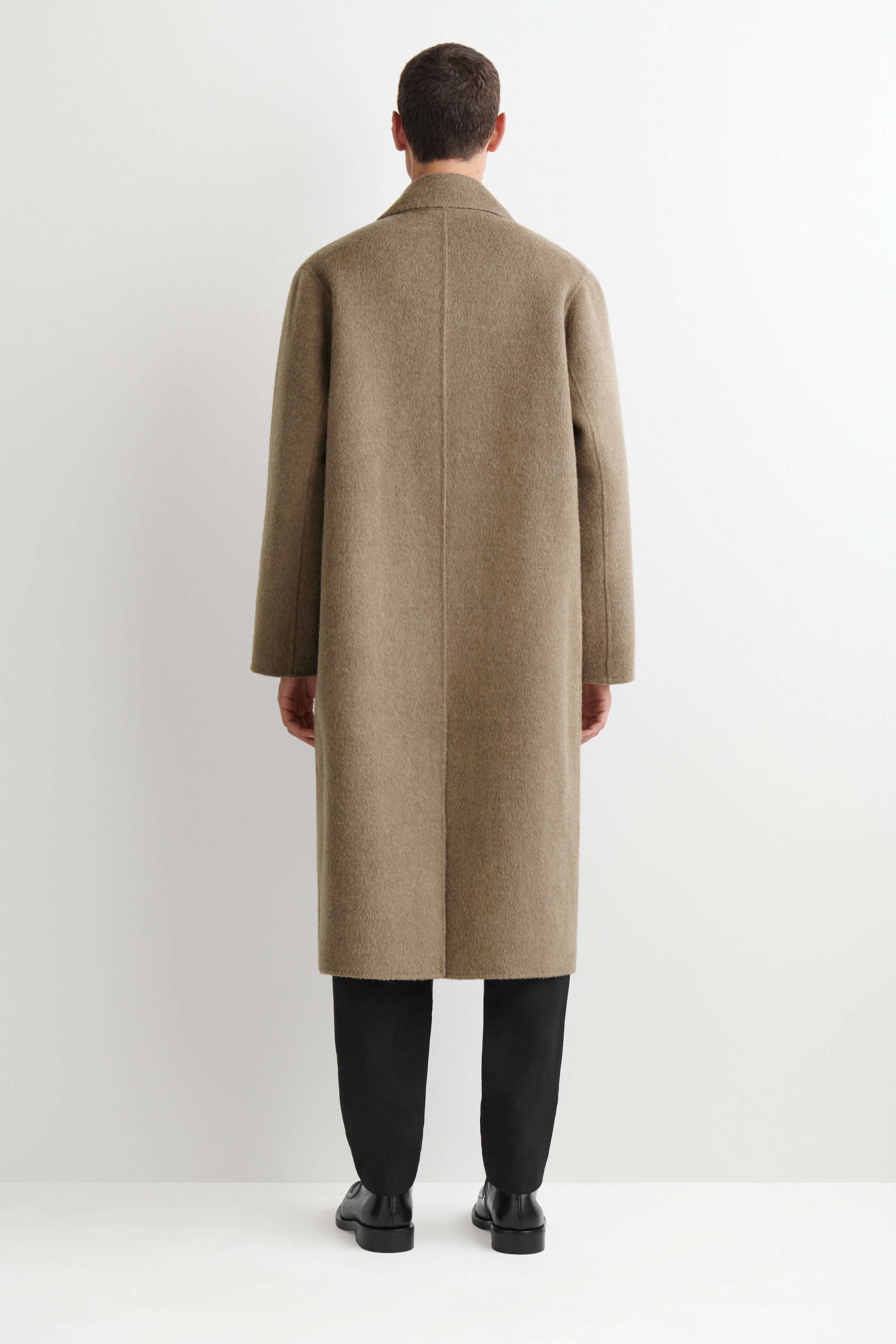 A double-faced wool-blend long coat in a taupe-brown shade is shown from the back, highlighting its straight-cut silhouette and clean center seam. The coat falls to mid-calf length, showcasing its soft, brushed texture and is styled with black trousers.