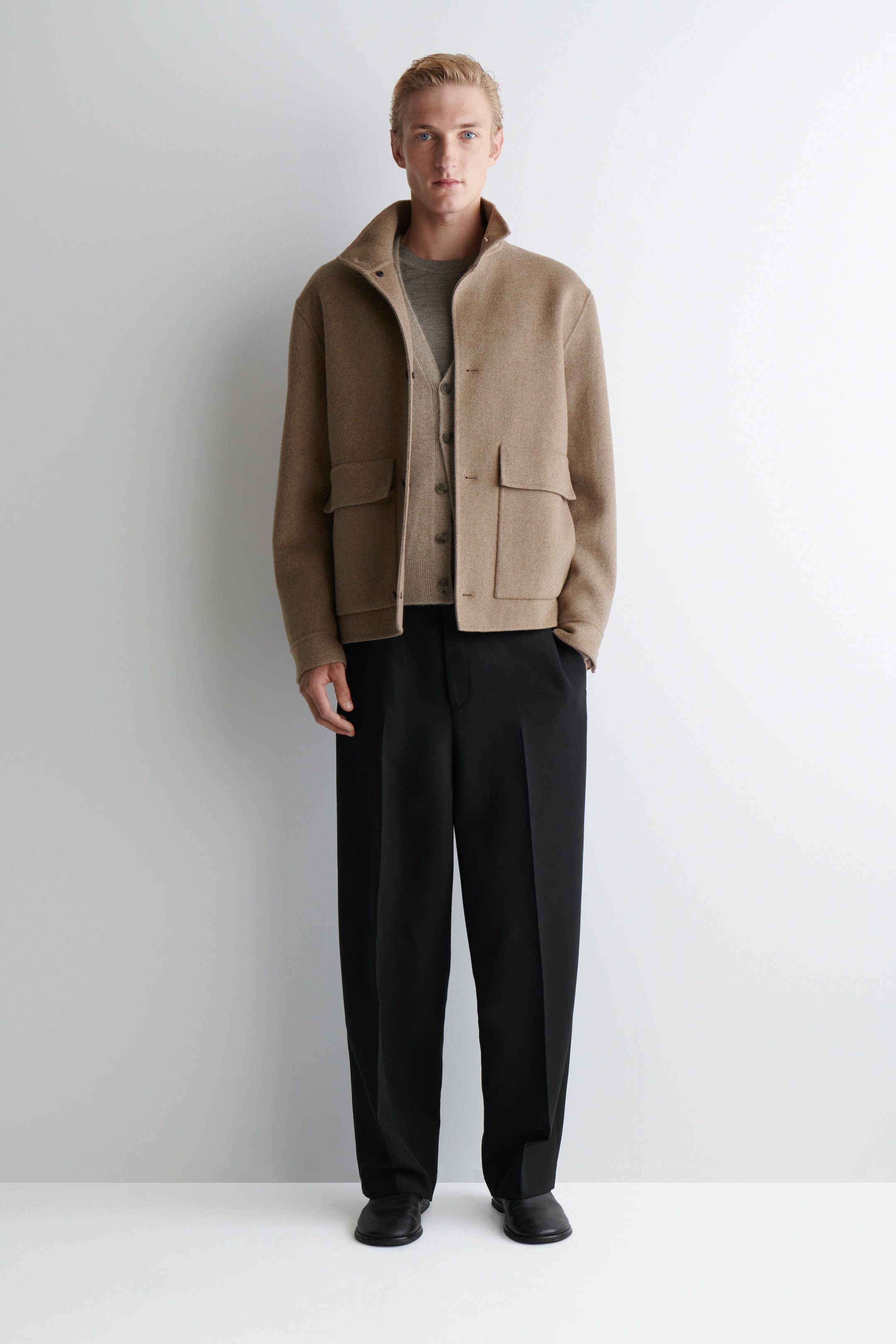 Grösseres Bild ansehen: Eine beige, strukturierte Jacke wird von vorne präsentiert, mit einem Stehkragen mit sichtbaren Druckknöpfen und zwei großen Klappentaschen auf der unteren Vorderseite. Sie wird offen getragen, wodurch ein hellerer Cardigan darunter sichtbar wird.