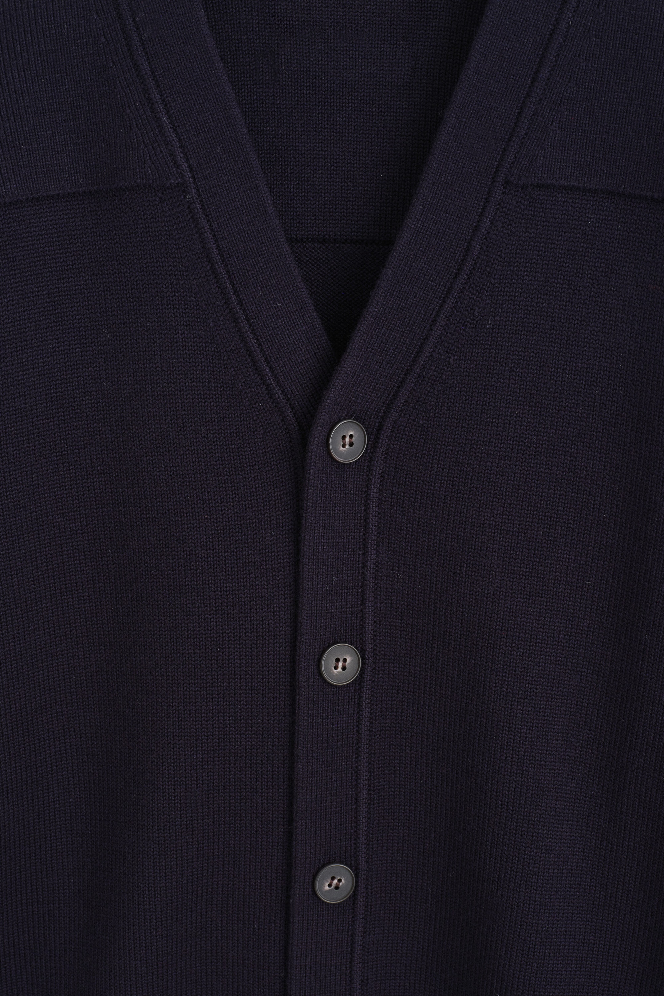 A navy blue cardigan features a V-neck and a button placket running down the centre front. Three dark, circular, four-hole buttons secure the placket, with horizontal panels visible on the upper chest. A navy blue cardigan features a V-neck and a button placket running down the centre front. Three dark, circular, four-hole buttons secure the placket, with horizontal panels visible on the upper chest.