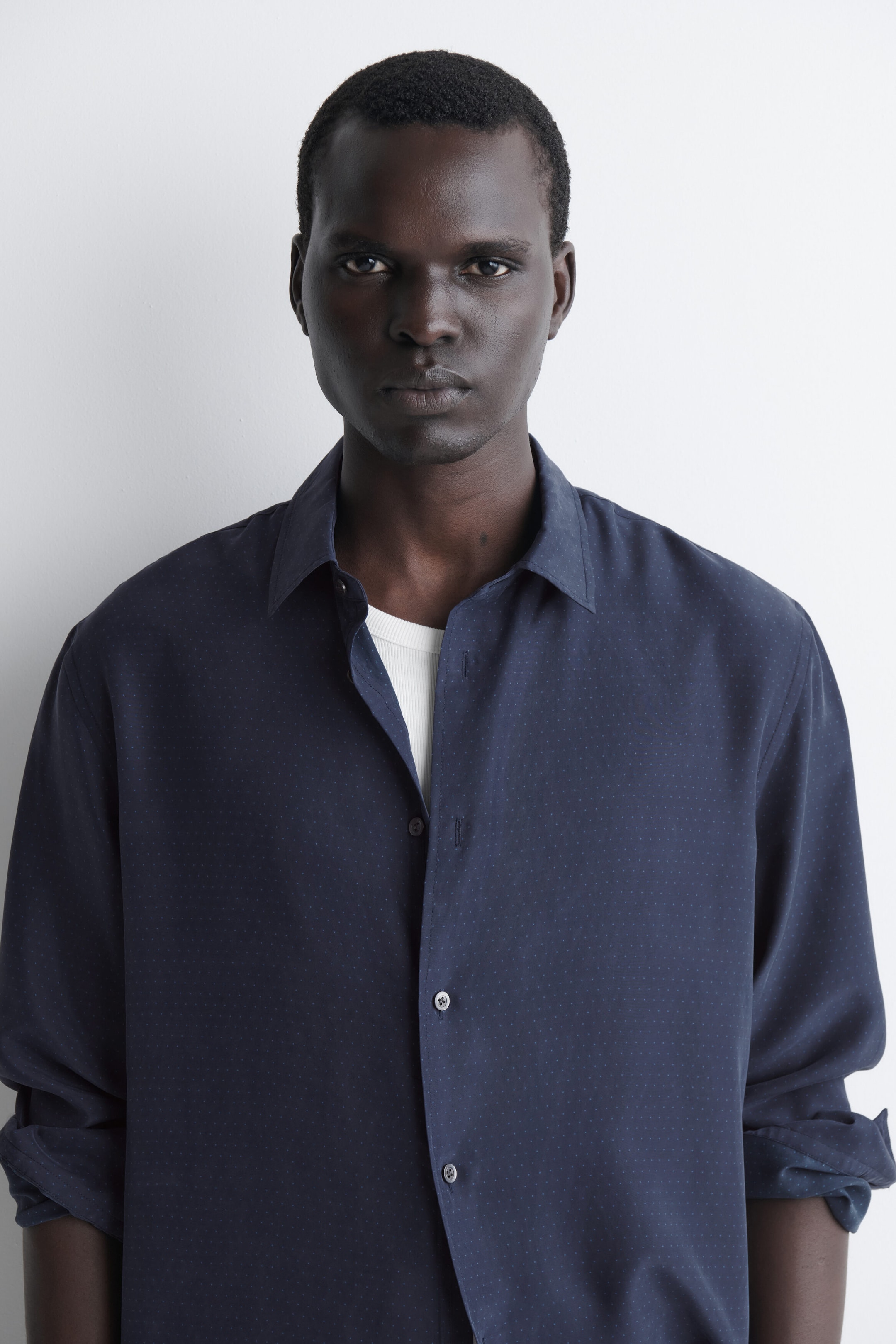 A navy blue sandwashed silk shirt with a subtle micro-dot pattern is shown from the front, draping in a relaxed fit. It is worn open at the collar over a white ribbed undershirt, with the sleeves rolled up to the forearms. A navy blue sandwashed silk shirt with a subtle micro-dot pattern is shown from the front, draping in a relaxed fit. It is worn open at the collar over a white ribbed undershirt, with the sleeves rolled up to the forearms.