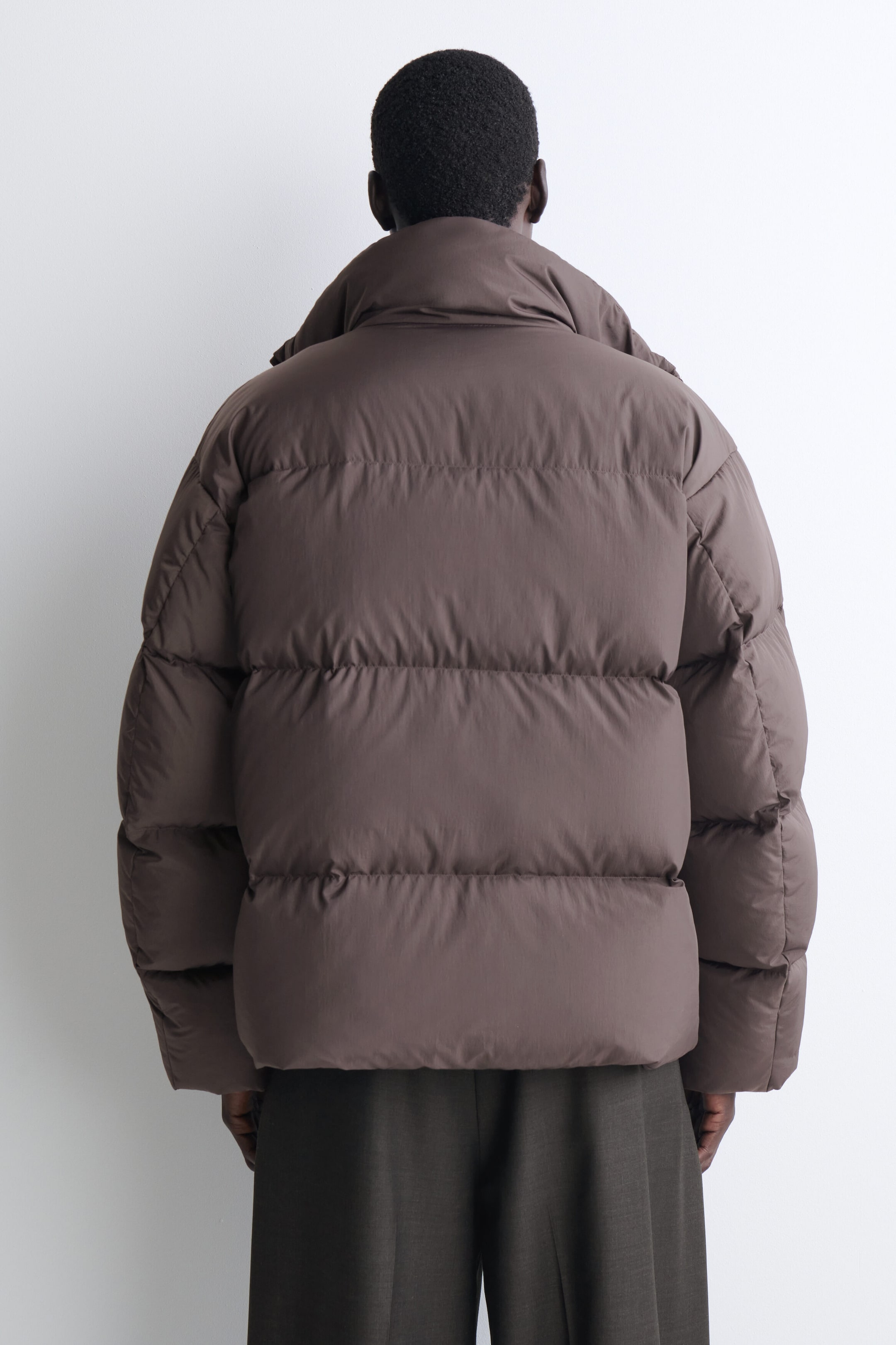 The funnel-neck nylon down puffer jacket is shown from the back, highlighting its voluminous, boxy fit and wide horizontal quilting in a muted brown tone. The jacket features a high, padded funnel collar and rests over dark trousers. The funnel-neck nylon down puffer jacket is shown from the back, highlighting its voluminous, boxy fit and wide horizontal quilting in a muted brown tone. The jacket features a high, padded funnel collar and rests over dark trousers.