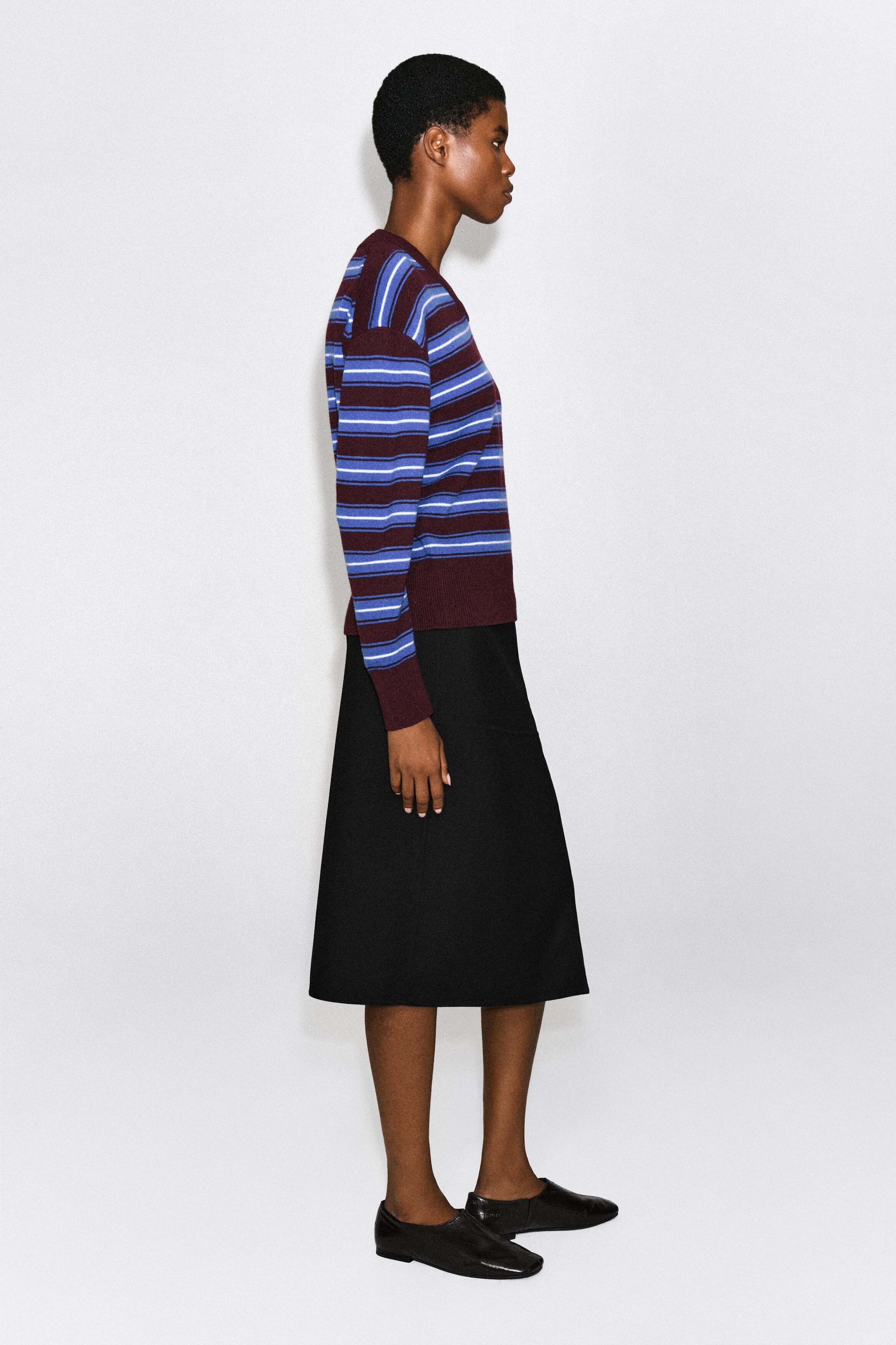 This merino wool layered V-neck sweater, featuring horizontal stripes in blue, burgundy, and white, is worn by the model standing in profile. The long-sleeved sweater is styled with a black midi skirt and black flats. This merino wool layered V-neck sweater, featuring horizontal stripes in blue, burgundy, and white, is worn by the model standing in profile. The long-sleeved sweater is styled with a black midi skirt and black flats.