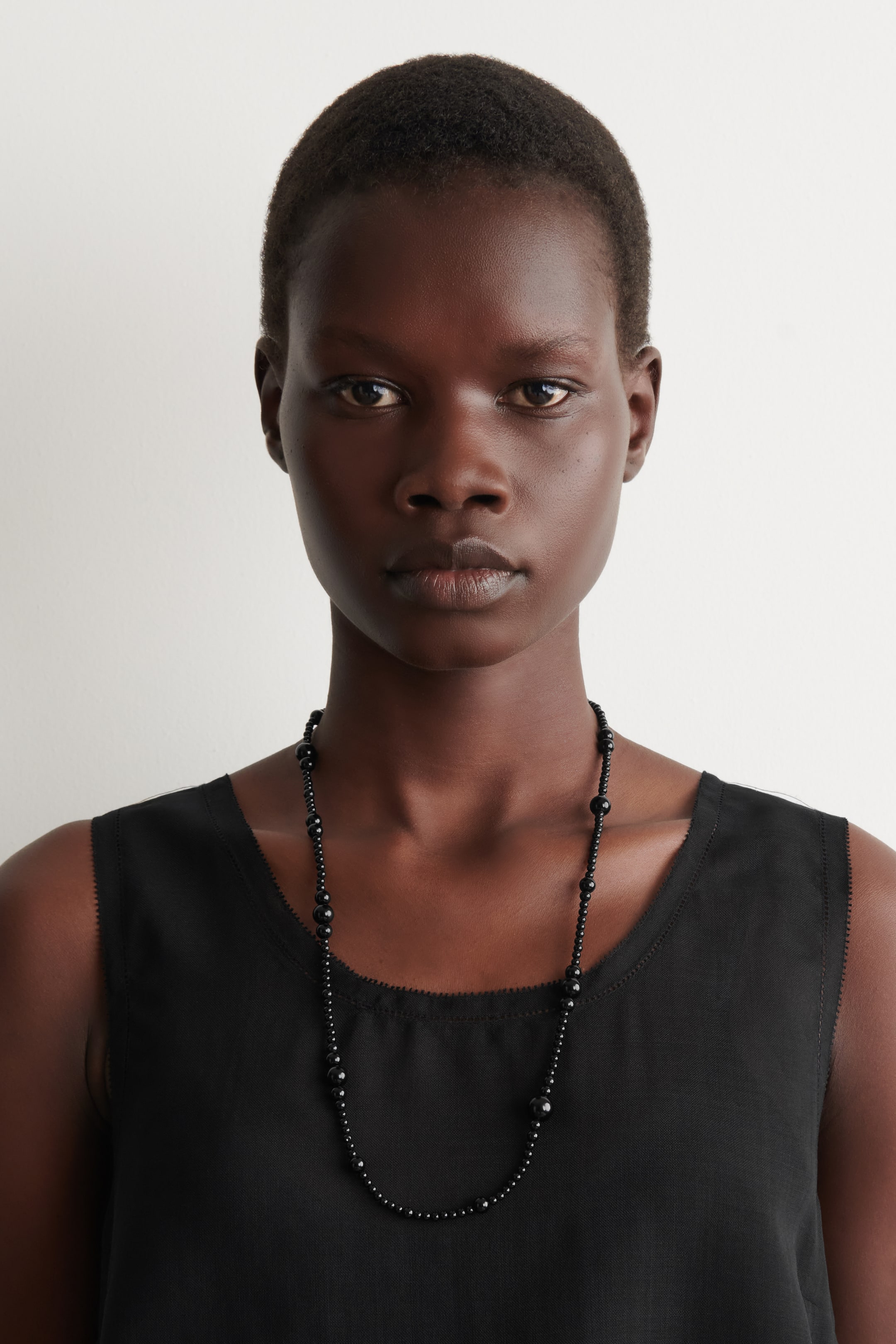 Ver imagen más grande: Un collar de cuentas negras presenta cuentas pequeñas y uniformes intercaladas con cuentas más grandes, redondas y pulidas, que se colocan alrededor del cuello y caen sobre la parte delantera de una blusa negra sin mangas.