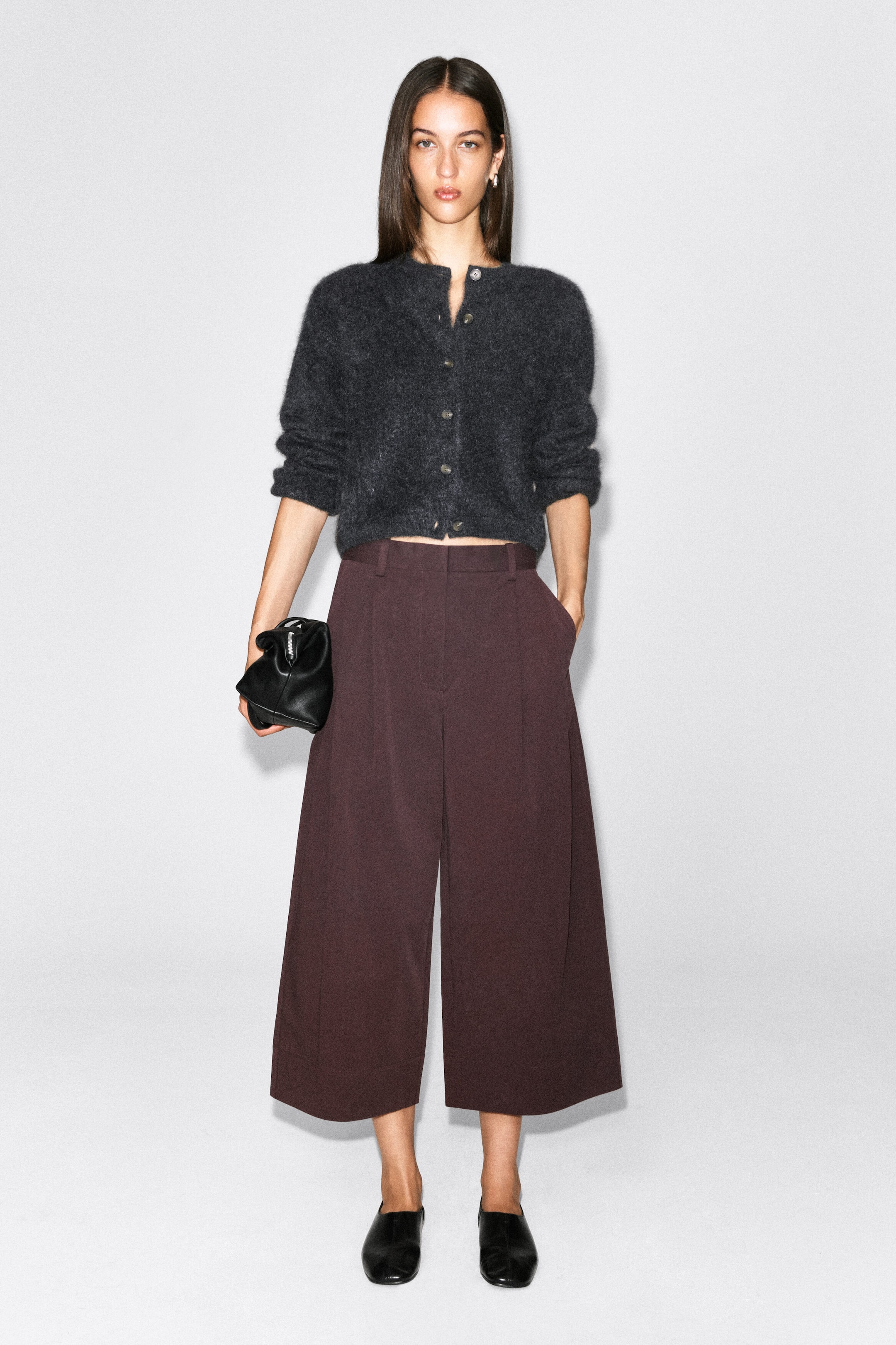 Cotton cropped barrel-leg pants in a deep burgundy shade are worn by the model, featuring a high-rise waist with belt loops, front pleats, and side pockets. The pants are styled with a dark gray knit cardigan, black leather mules, and a small black clutch bag. Cotton cropped barrel-leg pants in a deep burgundy shade are worn by the model, featuring a high-rise waist with belt loops, front pleats, and side pockets. The pants are styled with a dark gray knit cardigan, black leather mules, and a small black clutch bag.