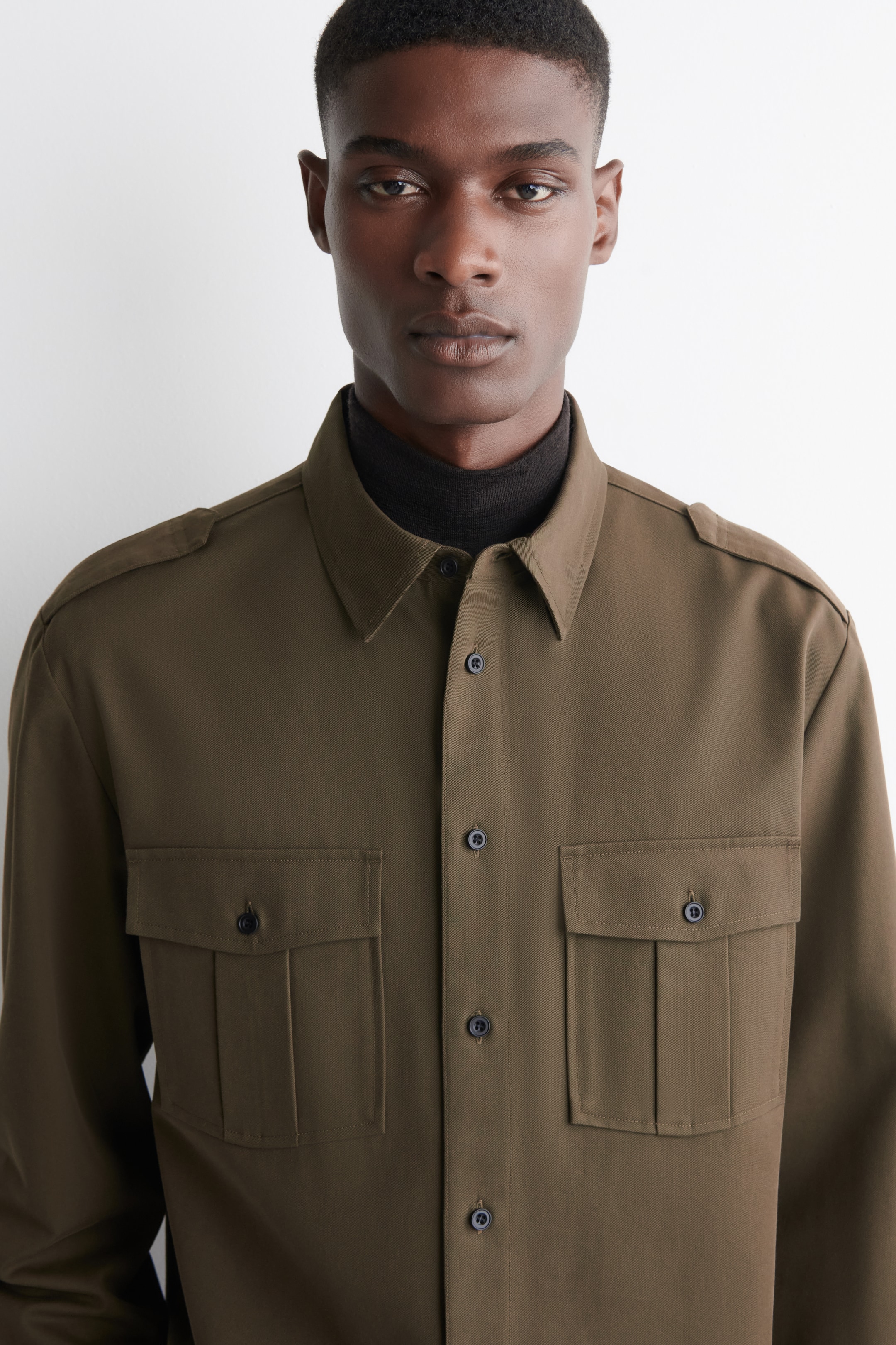 Agrandir l'image: Une chemise utilitaire vert olive est présentée de face, avec un col pointu, une fermeture complète à boutons avec des boutons sombres, des épaulettes et deux poches à rabat sur la poitrine avec des fermetures à boutons sombres et des plis creux. La chemise est superposée sur un col roulé côtelé foncé.