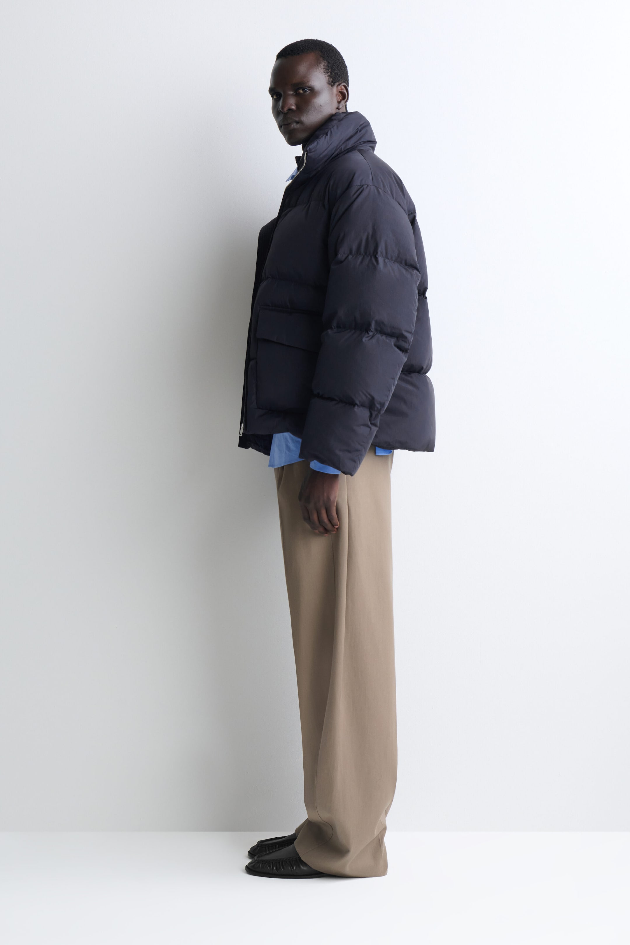 A navy funnel-neck nylon down puffer jacket is presented in side profile facing left, showcasing its voluminous silhouette and horizontal quilting. The jacket features a high stand collar and a large side flap pocket, and is styled over a light blue shirt and wide-leg beige trousers.