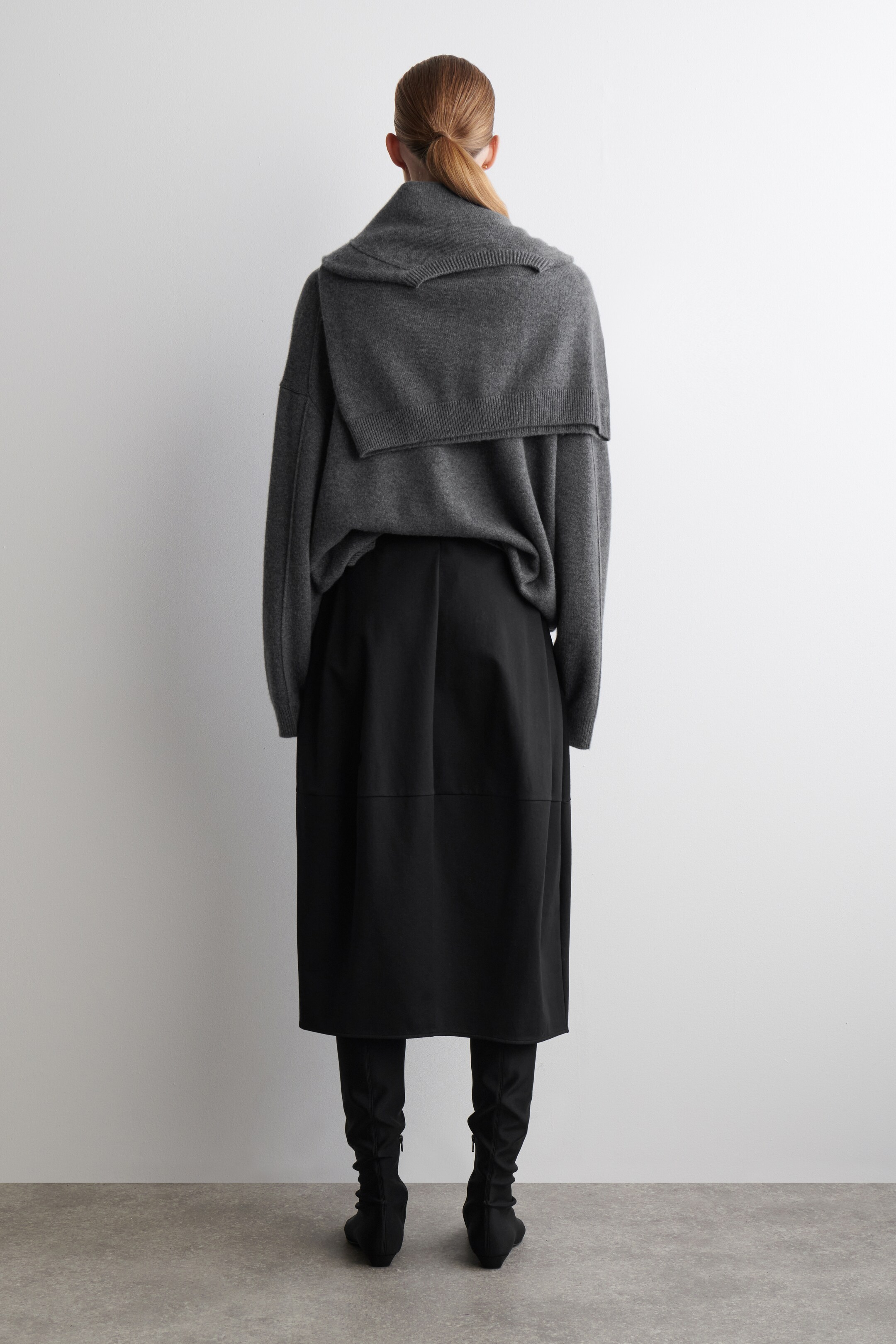 Agrandir l'image: Un pull en tricot gris foncé surdimensionné est visible de dos, mettant en valeur un large col châle côtelé qui tombe sur les épaules. Il est superposé à une jupe midi noire, qui présente un détail de couture horizontale, et des bottes noires jusqu'aux genoux, qui révèlent une fermeture éclair latérale subtile.