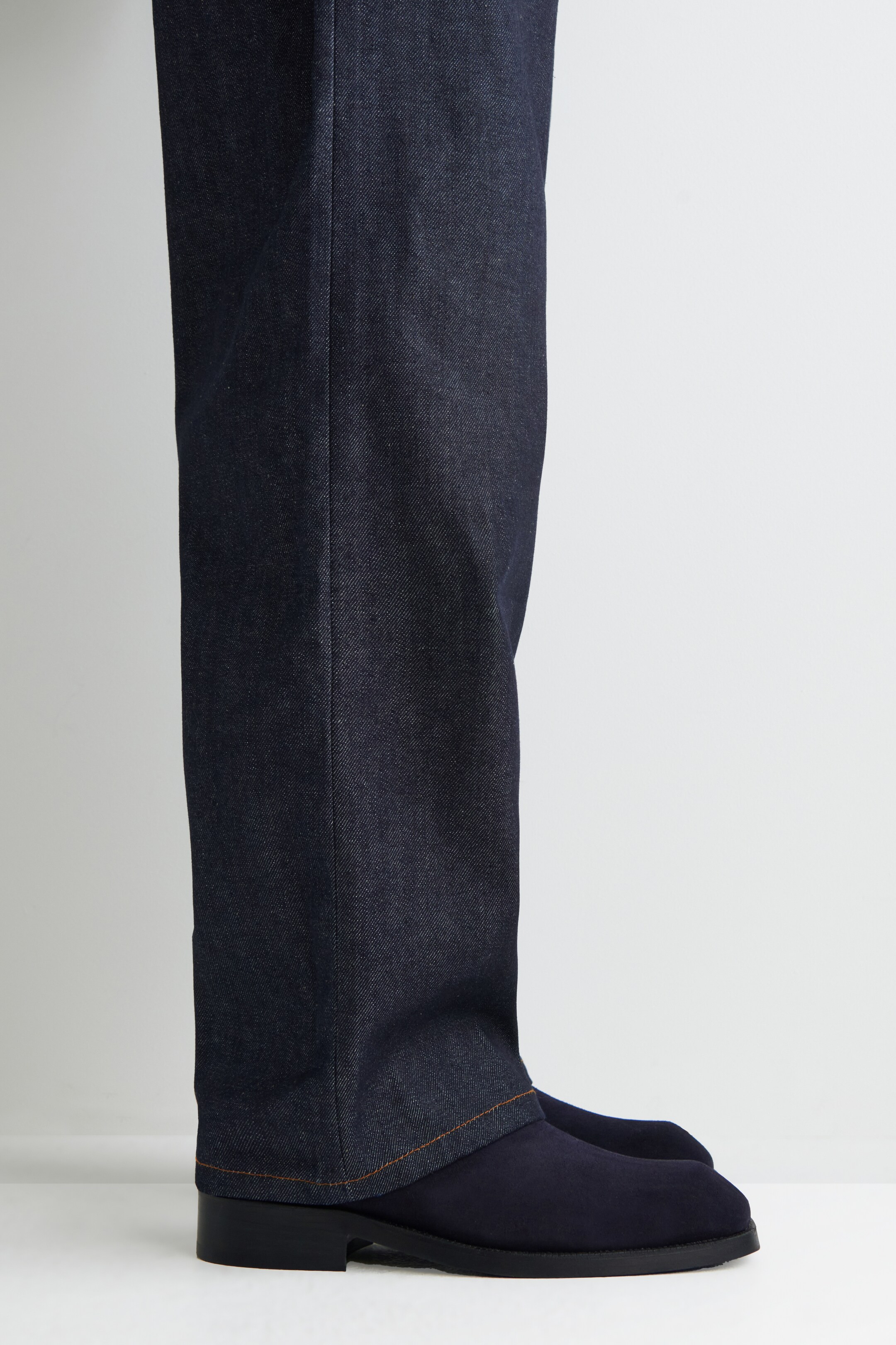 Grotere afbeelding bekijken: Donker indigo denim broek valt recht, met een zichtbare voorkant plooi en een oranje gestikte zoom. De zoom van de broek rust bovenop marineblauwe suède schoenen, die een lage zwarte hak tonen.
