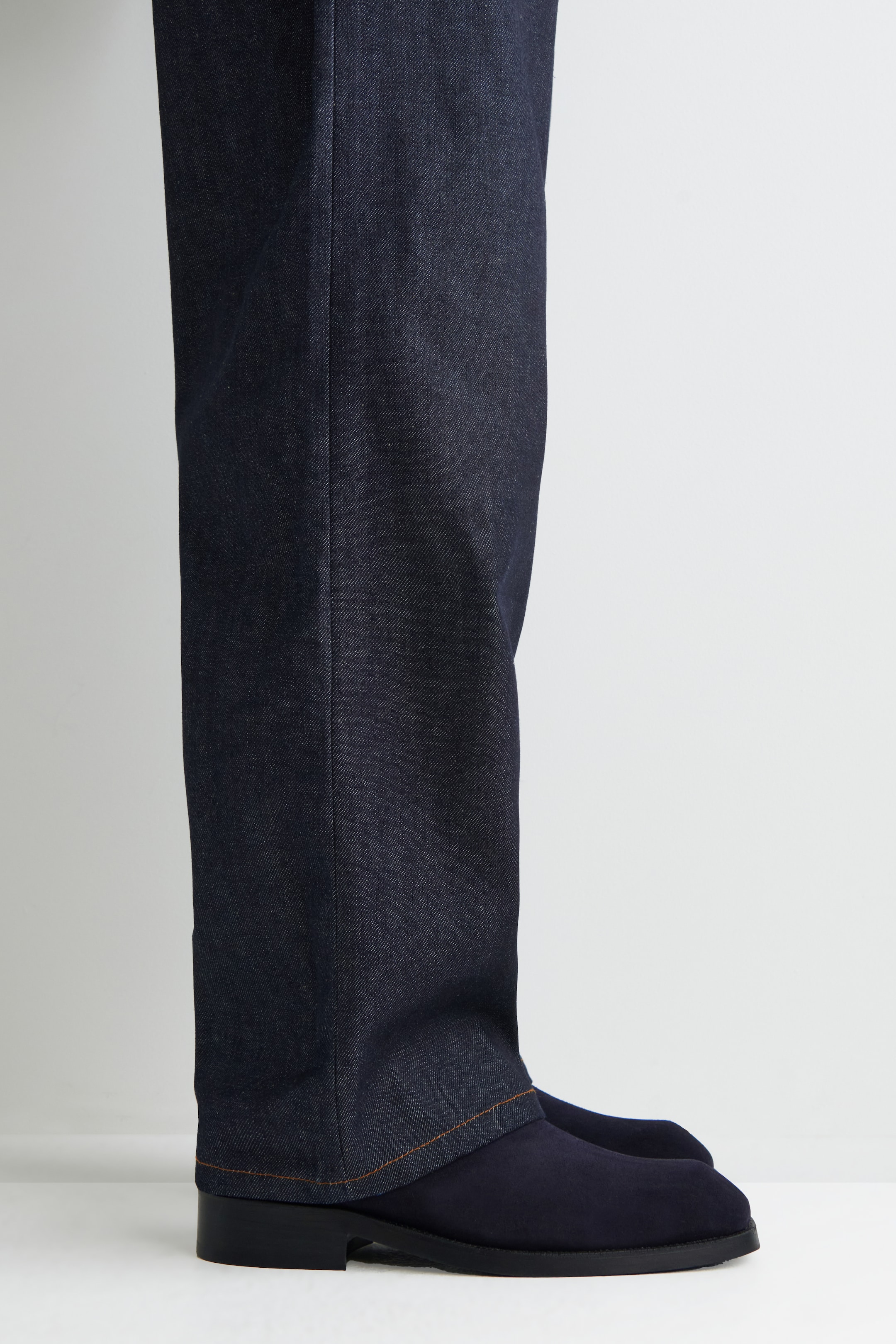 Agrandir l'image: Un pantalon en denim indigo foncé tombe droit, avec un pli visible à l'avant et un ourlet cousu en orange. L'ourlet du pantalon repose sur des chaussures en daim bleu marine, qui affichent un talon bas noir.