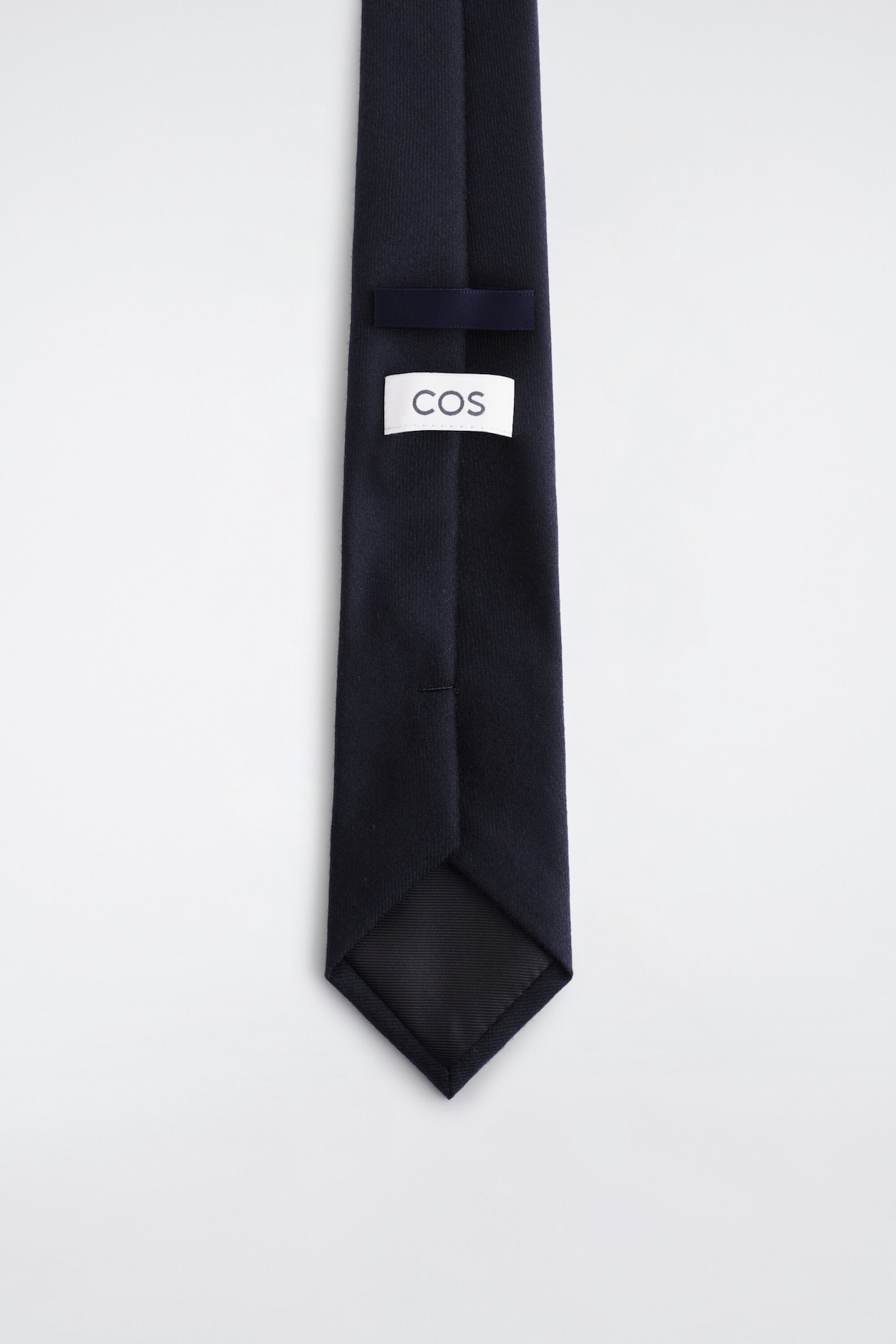 A navy blue necktie, shown from the back, displays a textured fabric construction with a matching fabric keeper loop and a white rectangular brand label featuring the text 'COS'. The pointed tip of the tie reveals a smooth, dark lining and a vertical centre seam.