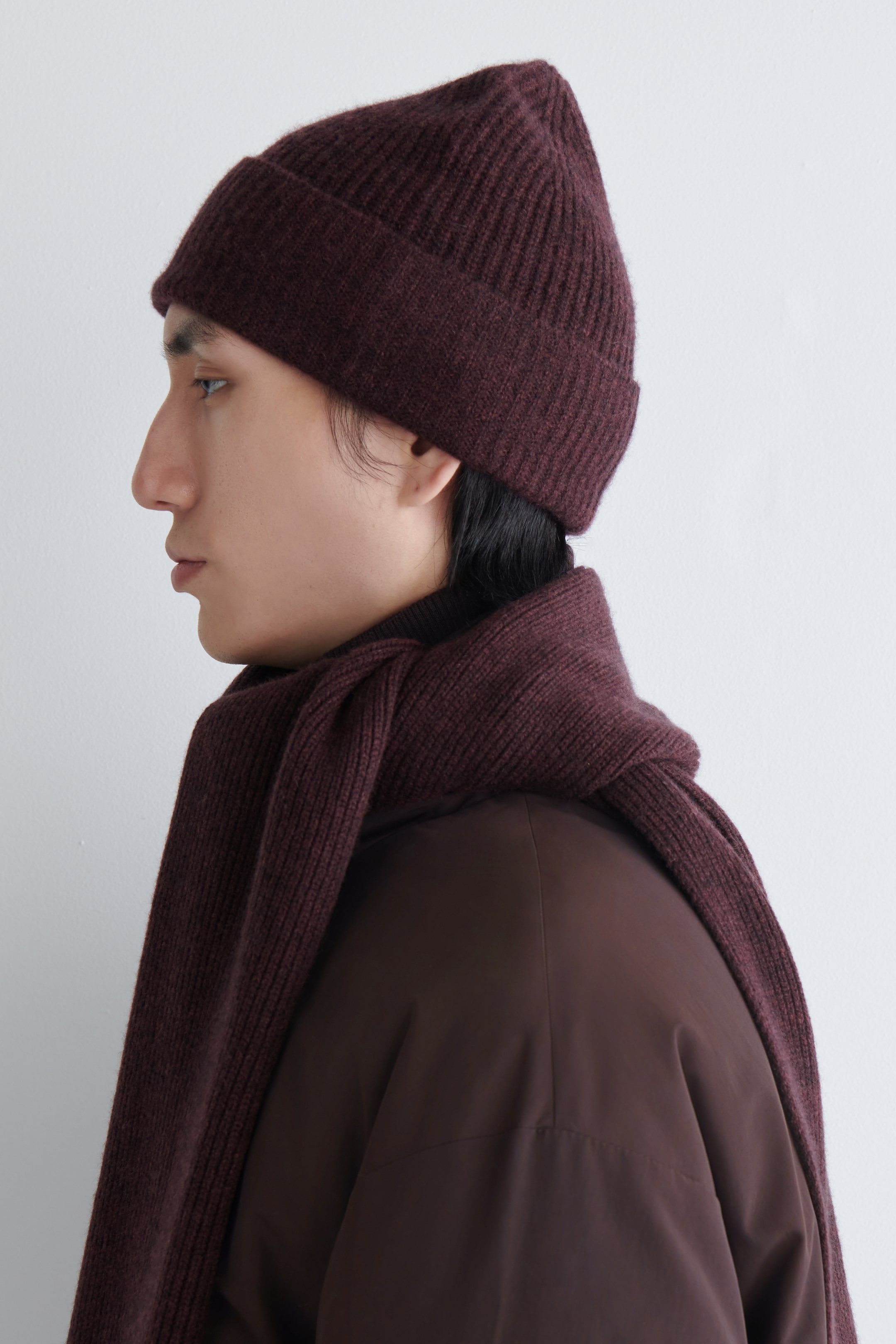 View larger image: A maroon rib-knit beanie is being worn on the model's head. A matching rib-knit scarf is being wrapped around the model's neck, layering over a dark brown jacket.