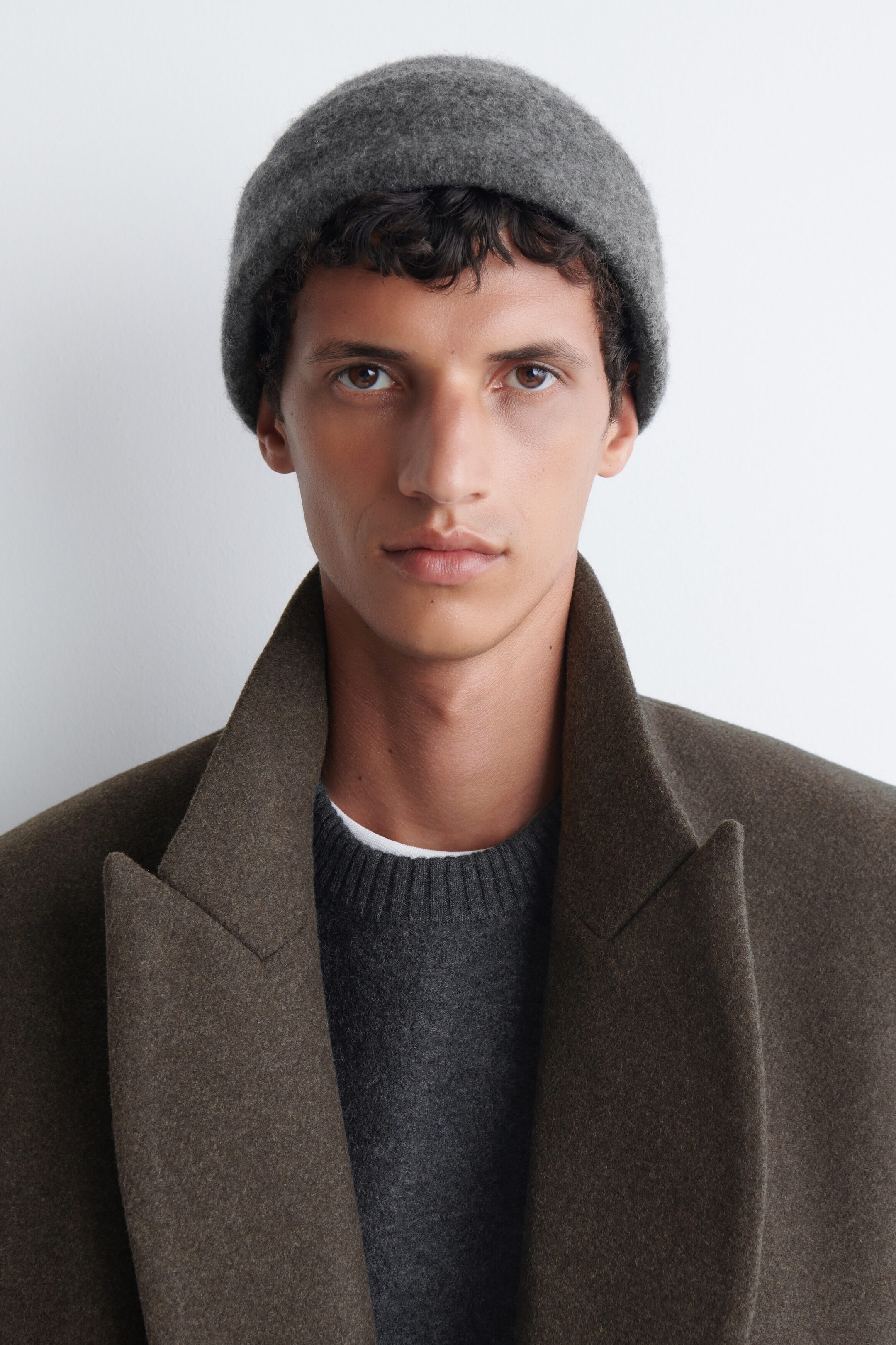 Un bonnet en laine gris avec revers est porté par le modèle, agencé avec un chandail à col rond gris foncé et un manteau en laine brun à larges revers. Un bonnet en laine gris avec revers est porté par le modèle, agencé avec un chandail à col rond gris foncé et un manteau en laine brun à larges revers.